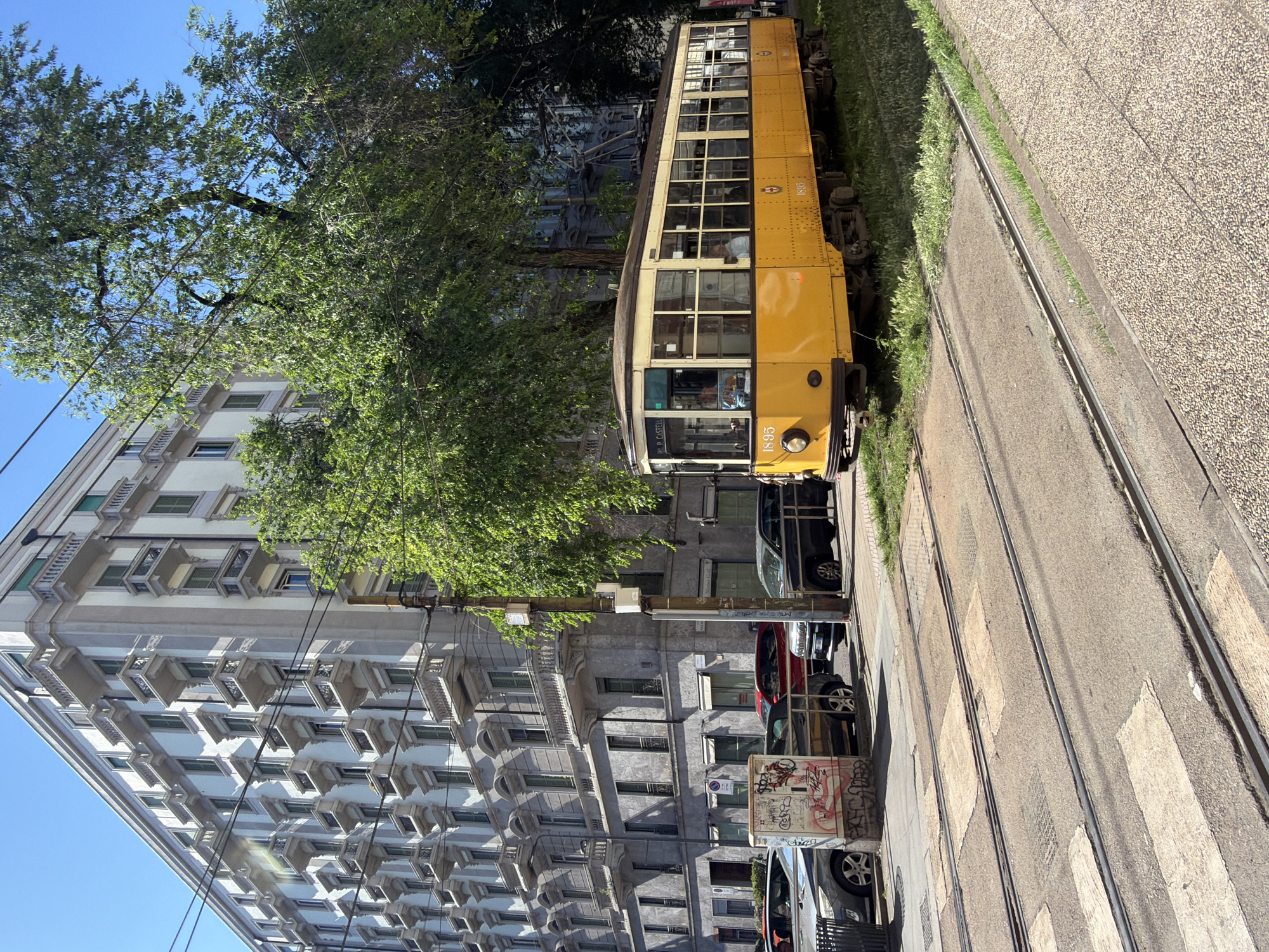 The image shows a street scene in Milan, Italy, featuring a vintage yellow tram. The tram is positioned on a street track running alongside a sidewalk. A tall, ornate building with multiple windows and balconies is situated next to the tram. The scene includes parked cars, a tree with green foliage that slightly obscures the building and tram, and overhead electrical wires. The background consists of a mix of sunlight and shadows.