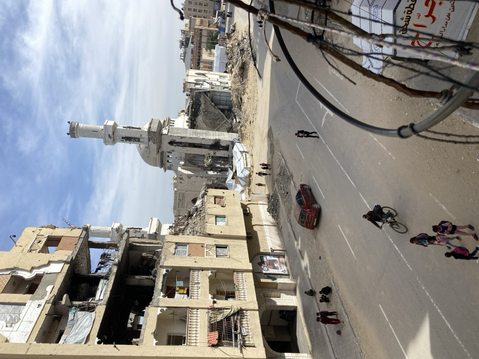The image displays a street in Khan Yunis, Palestinian Territory, under a partly cloudy sky, characterized by extensive urban destruction. On the left, a multi-story building is severely damaged, with large sections of its facade missing, revealing exposed interiors and rebar; laundry hangs from a third-story balcony. In the mid-ground, a large mosque with a prominent minaret and dome is significantly damaged, its surrounding area reduced to substantial piles of rubble and debris. White temporary structures are visible beyond the mosque.
The paved street in the foreground features white double solid lines. A red sedan is parked facing left on the right side of the road. Multiple individuals are present: on the lower left, approximately four children are gathered on the sidewalk near a deflated ball; a man cycles towards the viewer in the center; a group of four children, some with backpacks, walks away from the viewer below the cyclist; and on the right side of the road, a man carries a small child while walking towards the viewer. Additional smaller figures are visible walking across the rubble-strewn terrain in the background near the mosque.