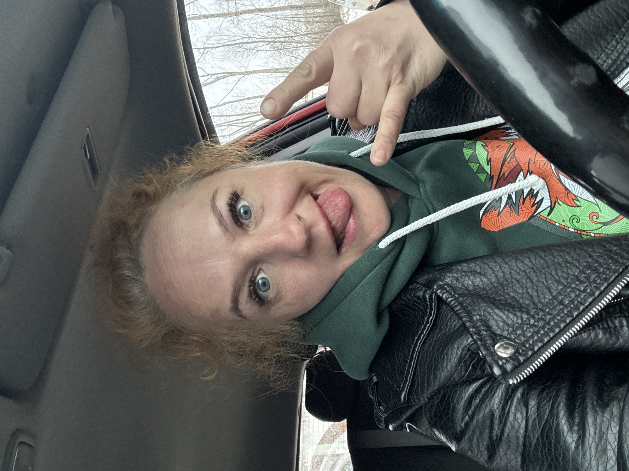 A woman with blonde curly hair and blue eyes is in the driver's seat of a car, sticking her tongue out. She's wearing a green hoodie with a colorful design on the front and a black leather jacket. Her left hand is visible, making a "rock on" hand gesture. The steering wheel of the car is visible in the upper right of the frame. The background shows trees, suggesting an outdoor setting, possibly in the city of Chaikovsky, Russia. The car's interior is visible, including the sun visor.