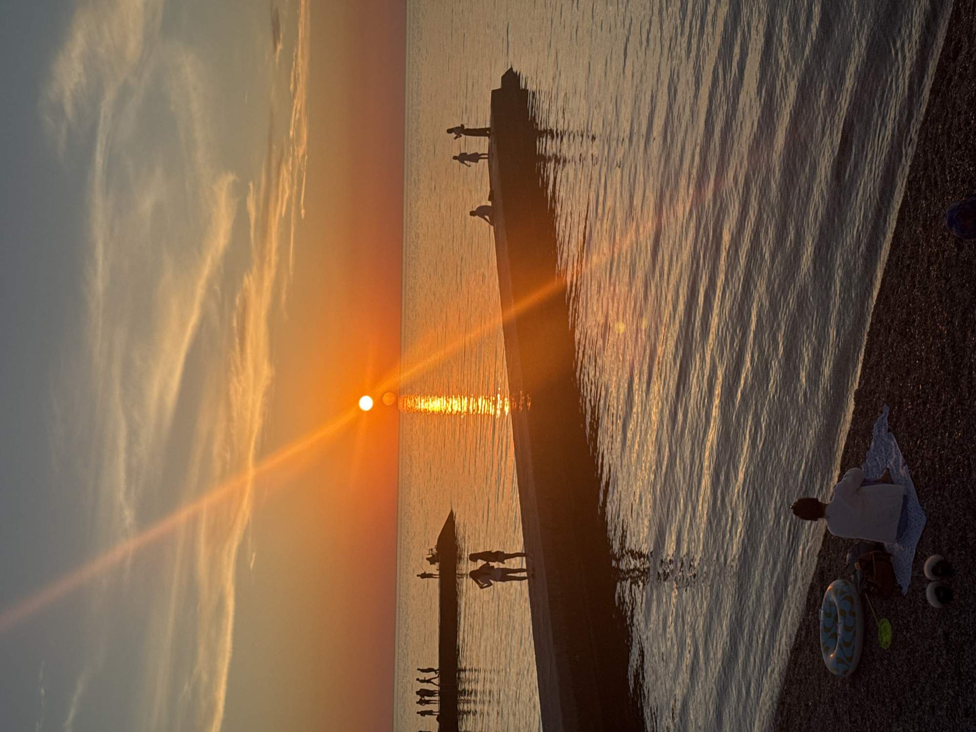 This is a vertical, outdoor, eye-level, medium shot of a beach scene at sunset. The sun is low on the horizon, casting a warm orange and yellow glow across the sky and water, with a strong sun flare visible across the image. The sky is a gradient of blue and orange, with thin, wispy clouds.
In the foreground, a person sits on a towel on a pebble beach, facing away from the camera and towards the water. Next to them are an inflatable ring, a beach bag, a toy shovel, and a pair of fuzzy slippers.
In the middle ground, two piers extend into the calm, reflective water. On the closer pier, two young people stand near the edge, facing each other. Further out on this pier, a person is seated. On a second pier in the background, a small group of people are visible, some standing and some walking. One person on this pier appears to be fishing. The water is a deep blue-gray, with subtle ripples reflecting the colors of the sunset.