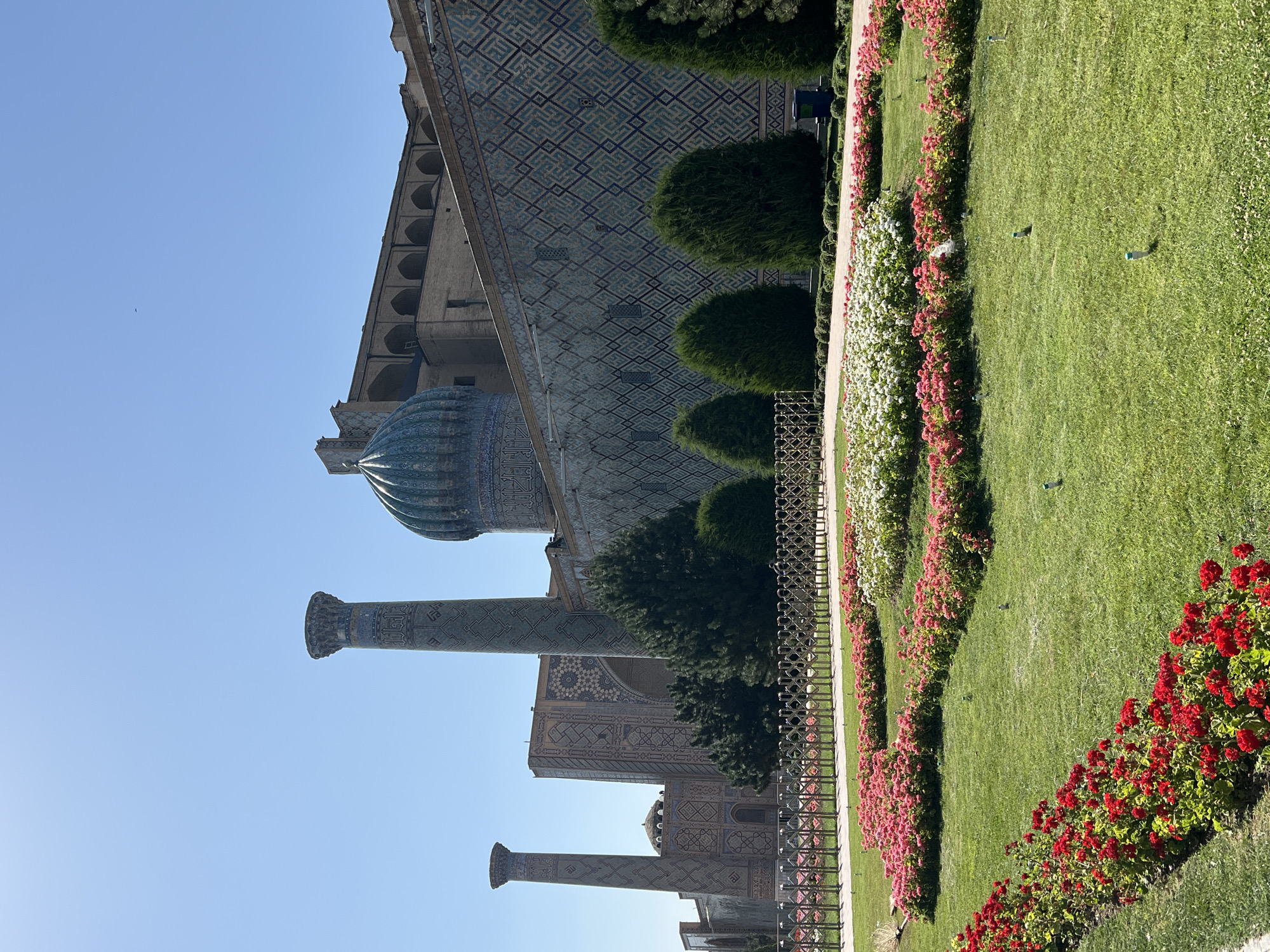 The image shows a view of the Registan Square in Samarkand, Uzbekistan, on a clear, sunny day. The foreground features a meticulously maintained lawn with several beds of colorful flowers, primarily red and pink, arranged in curved patterns. Several white and pink flowers are also visible in clusters further back. Sprinklers are dotted throughout the lawn. Behind the flowerbeds and lawn is a low, decorative metal fence.
Beyond the fence, the imposing Registan complex dominates the scene. Two tall, ornate minarets, adorned with intricate blue and white tilework, rise prominently against the sky. One minaret is closer to the viewer and more to the left, while the other is further back and to the left of the first. Between and behind these minarets, the complex's facades are visible, featuring extensive geometric patterns in blue and white tiles. A large, turquoise-tiled dome of a madrasah is visible, as well as arched windows and the weathered brickwork of the main structures. Several mature, dark green trees are planted in a line behind the fence and in front of the buildings, adding depth to the composition. The sky is a clear, bright blue with no clouds. No people are clearly discernible in the image.