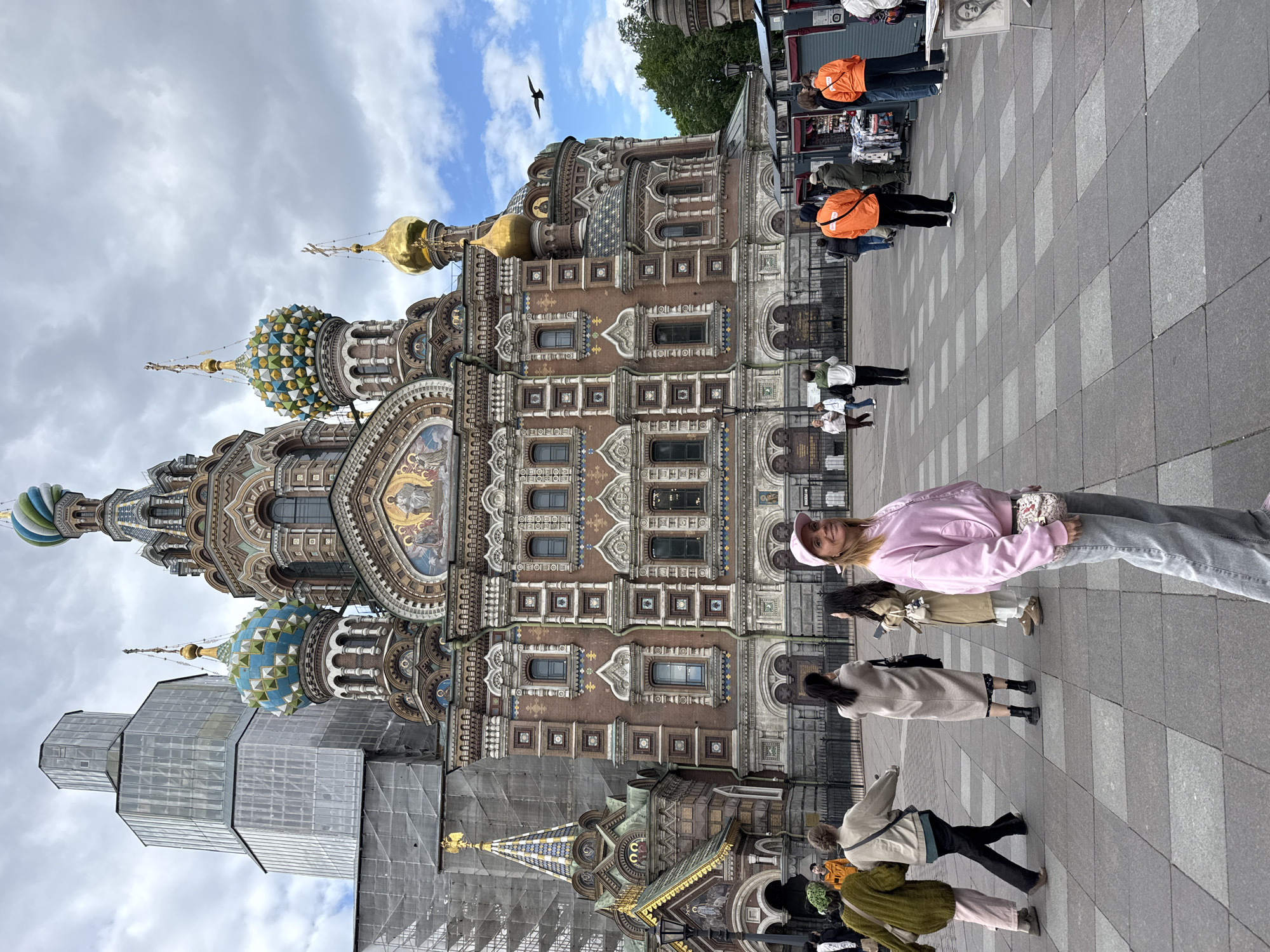 The image shows the Church of the Savior on Spilled Blood in Saint Petersburg, Russia, taken on a cloudy day. The church's intricate architecture and colorful domes are visible, with parts of it covered in scaffolding. A black bird flies in the sky above the church. A group of people are walking on the pavement in front of the church, including a woman in a pink jacket and hat. Some people are browsing goods displayed on tables.