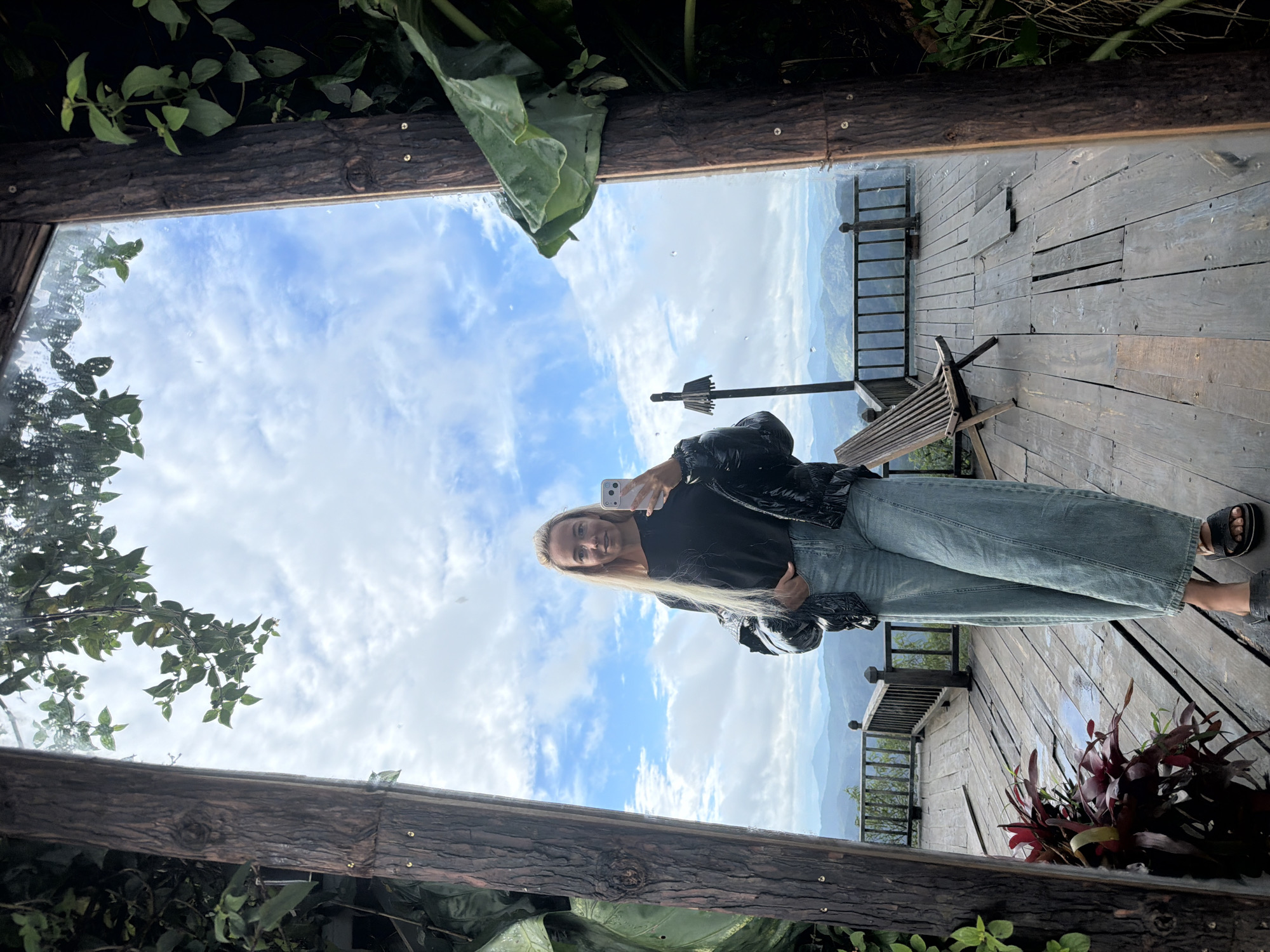 A full-body, front-facing selfie is taken by a blonde woman standing on a wooden deck. She is wearing a black long-sleeved shirt, a black puffer jacket, light blue wide-leg jeans, and black sandals. She holds a white smartphone in her right hand to take the photo.
The wooden deck is elevated and overlooks a mountainous landscape. The background features a hazy, blue sky with scattered clouds, and rolling hills receding into the distance. A wooden railing lines the edge of the deck. To the left of the woman, there is a dark-colored potted plant with red leaves. To her right, a weathered wooden lounge chair is positioned on the deck.
The scene appears to be outdoors, possibly at a viewpoint or a resort in a mountainous region. The presence of the wooden deck, railing, and lounge chair suggests a public or semi-public outdoor space designed for relaxation and enjoying the view. The natural elements, such as the trees and foliage framing the mirror, and the mountainous scenery, point towards a location outside of a dense urban area. The overall atmosphere is tranquil and scenic.
It is not possible to definitively identify the location as being specifically within the city of Phát Chi, Vietnam, based solely on the visual information provided in the image.
