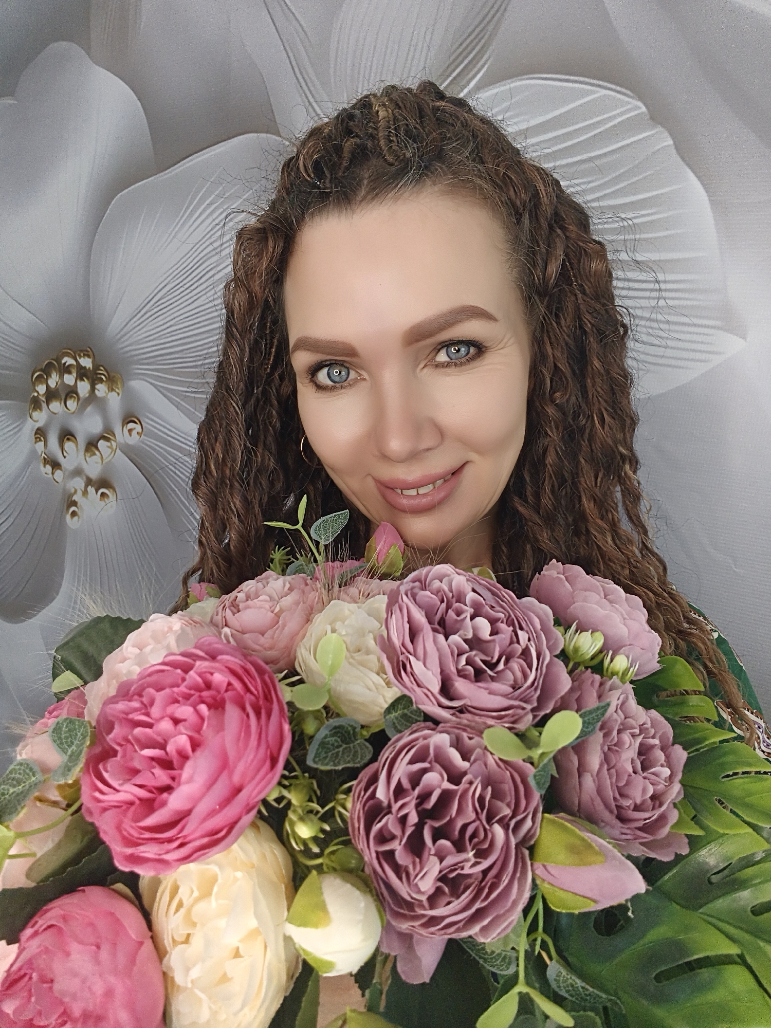 A woman with long curly brown hair is holding a bouquet of pink and cream-colored flowers in front of a white flower-patterned background. She has blue eyes and is smiling. The bouquet includes peonies and greenery. The background features a large white flower with a textured petal design. There's also another smaller white flower in the background with a central detail resembling gold-colored beads. The location is in the city of Unknown, Unknown.