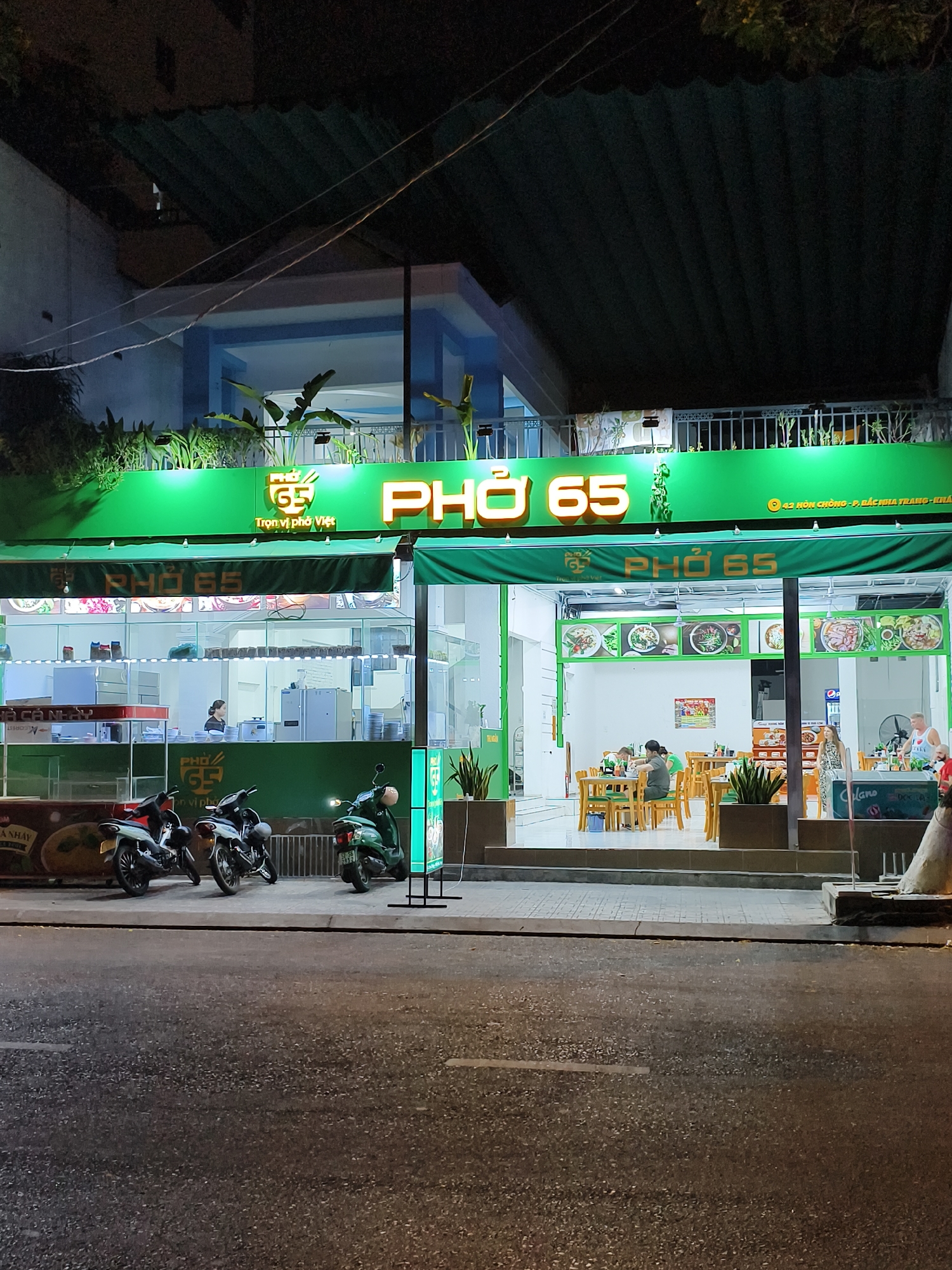 The image captures the exterior of "Phở 65," a Vietnamese restaurant located at 42 Hòn Chồng, in the Bách Nha Trang Ward of Nha Trang, Vietnam. It's nighttime. The restaurant's facade is prominently green, with bright signage illuminating "PHỞ 65." Inside, people are seated at wooden tables and chairs, seemingly dining. A woman stands behind a counter near the front. Three motorcycles are parked along the curb in front of the restaurant. The street in front of the restaurant appears wet.