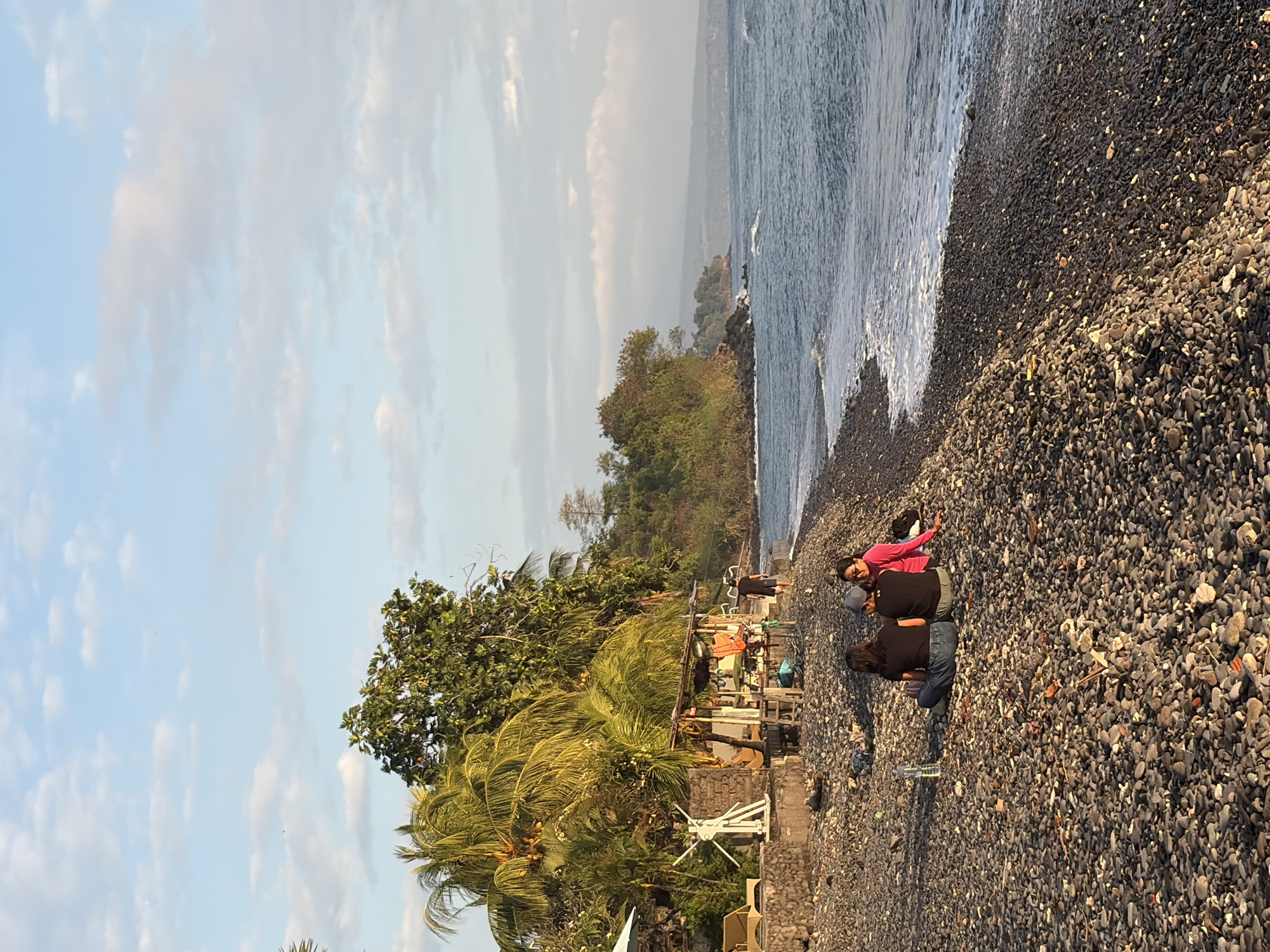 This is a vertical, eye-level, medium shot of a pebble beach with a line of trees and a small building on the left, and the ocean on the right. Three people are sitting on the pebbles in the foreground, with their backs to the camera, facing the ocean. The person on the left is wearing a black t-shirt and blue jeans. The middle person is wearing a black t-shirt and grey cap. The person on the right is wearing a bright pink long-sleeved shirt and sunglasses. A plastic water bottle sits on the pebbles to the left of the people.
Behind the people, there is a small, open-air structure with a thatched roof, propped up on stilts, with various items hanging from it, including orange cloths. To the left of the structure, there is a white, stylized sculpture on a pedestal. Further left, there are lounge chairs partially visible. A wall made of stone separates the beach area from the structures.
Palm trees and other lush green trees form a dense backdrop behind the structures and continue along the coastline. The ocean is a deep blue with gentle waves lapping the shore. The sky above is a pale blue with scattered white clouds. The overall lighting suggests it is late afternoon or early evening. The location is identified as Abang, Indonesia, based on the provided information.
