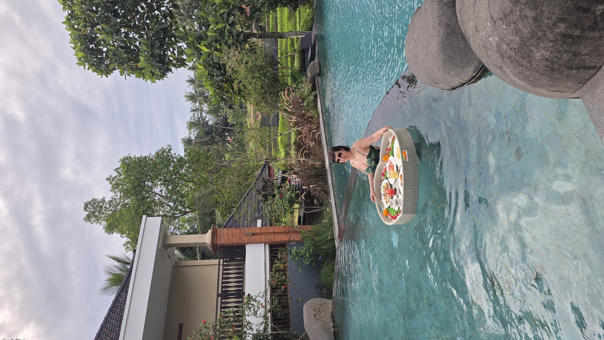 A medium shot depicts a woman in a turquoise swimming pool in Ubud District, Indonesia. She holds a round, floating tray laden with food and drinks. The pool is surrounded by vegetation, including trees and tall grasses. Behind the pool is a building with a white exterior and a tiled roof. The sky is overcast.
