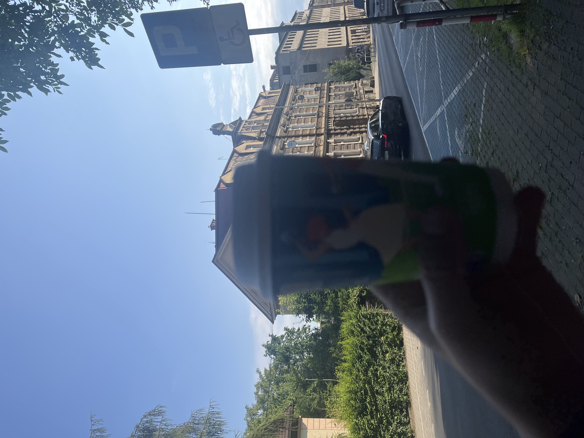 A person's hand holds a colorful paper coffee cup in the foreground, obscuring part of the view. Behind the cup, a street in a European city is visible. The street is lined with parked cars and buildings. One building, a multi-story structure with ornate architectural details, appears to be older. Another building, a more modern concrete structure, stands next to it. A blue parking sign with a white 'P' and a wheelchair symbol is mounted on a pole on the right side of the street. A black sedan is driving on the street. The sky is clear and blue, with some clouds visible. Green trees and hedges line the side of the street. The pavement is made of cobblestones.