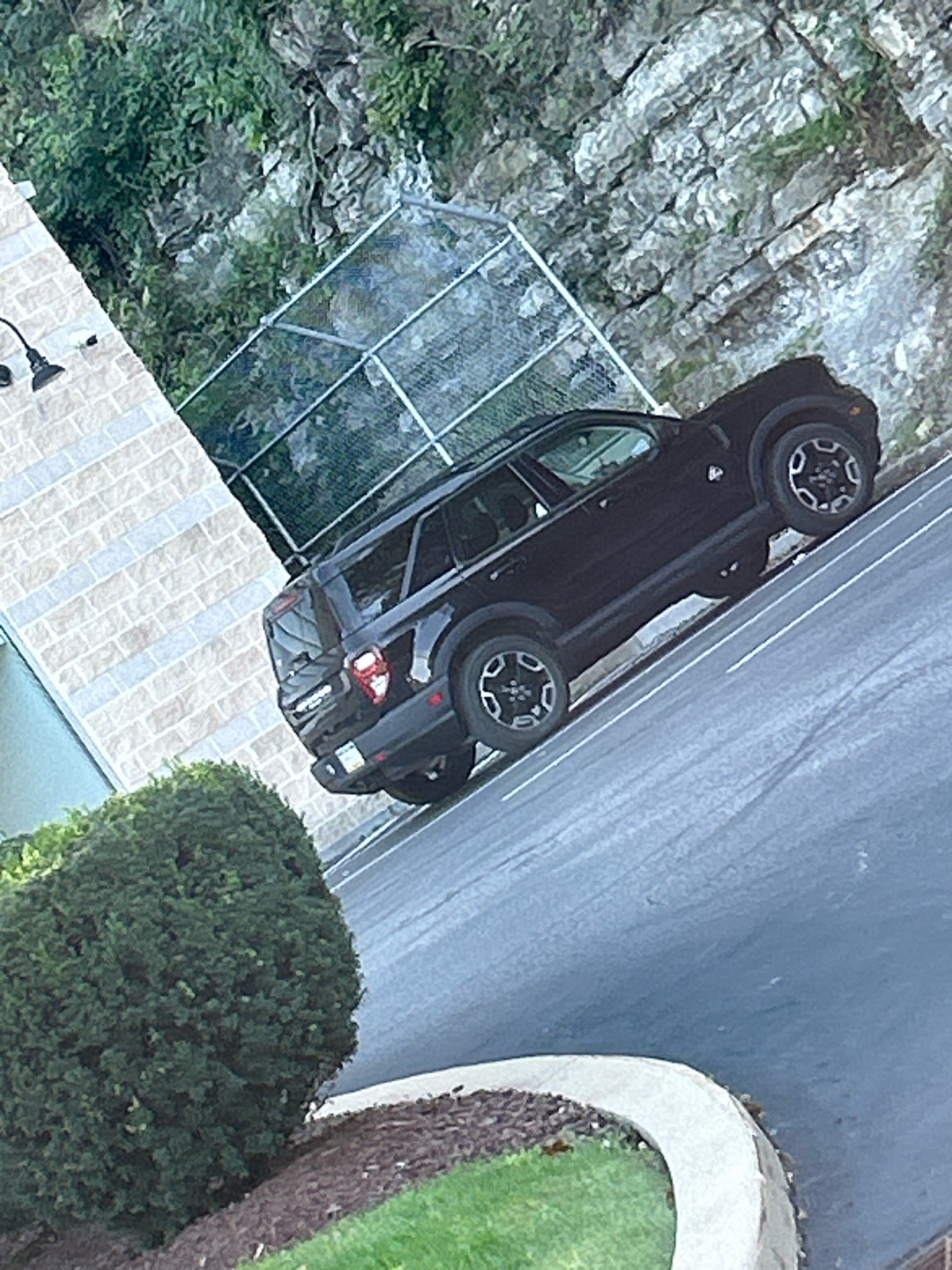 A black SUV is parked diagonally on a paved surface, possibly a parking lot or driveway, which slopes downwards. The vehicle is viewed from a high angle, tilted to the right. Behind the vehicle, a rocky cliff face covered in greenery rises sharply. To the left of the vehicle is a building with a light-colored brick facade, and a black outdoor light fixture is visible on the building. In the foreground, a neatly trimmed green hedge is situated next to a landscaped area with mulch and grass. A chain-link fence is positioned behind the vehicle, running diagonally across the frame. The scene appears to be outdoors during the daytime. The location is the city of Mt. Lebanon, United States of America.