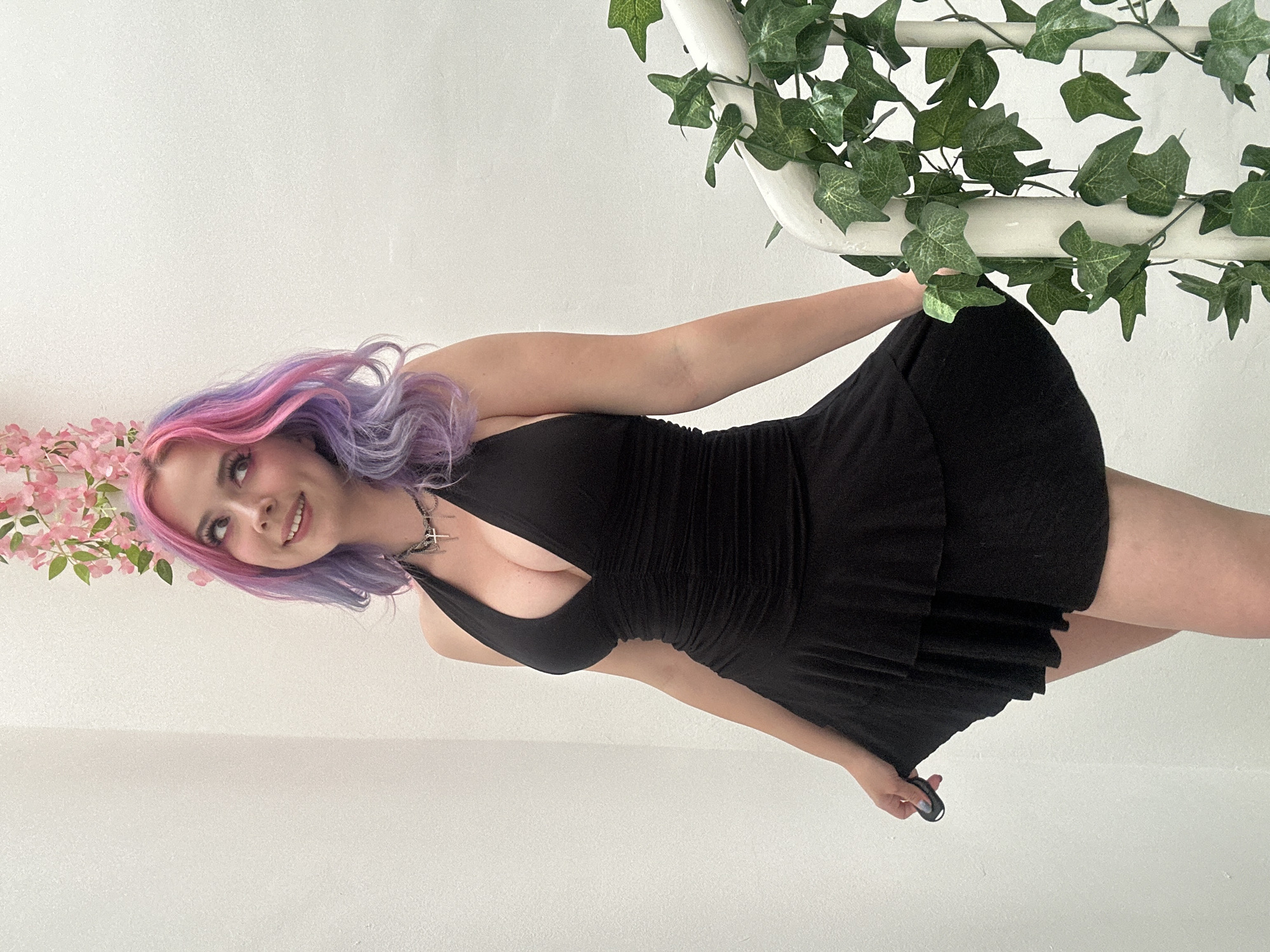 A young woman with long, wavy hair, dyed in vibrant shades of pink and purple, is depicted posing indoors in the City of Zagreb, Croatia. She is wearing a sleeveless black halter-neck mini-dress that gathers at the waist. A dark choker with a distinctive cross-shaped pendant adorns her neck. Her light-colored fingernails are visible, possibly painted blue or purple.
The woman is positioned diagonally across the frame, leaning back slightly with a gentle smile on her face, looking upwards and to her right. Her left arm is extended upwards, partially obscured by the foliage, while her right hand is subtly holding the hem of her dress near her right thigh.
The setting features a plain white wall as the background, creating a bright and clean ambiance. On the left side of the frame, near her head, are artificial pink cherry blossoms, while on the right, artificial green ivy is wrapped around a white, curved structure, possibly a railing or part of a spiral staircase. The lighting appears bright and even, suggesting daytime conditions indoors. There is no visible text in the image. The overall mood conveyed by her expression is pleasant and calm.