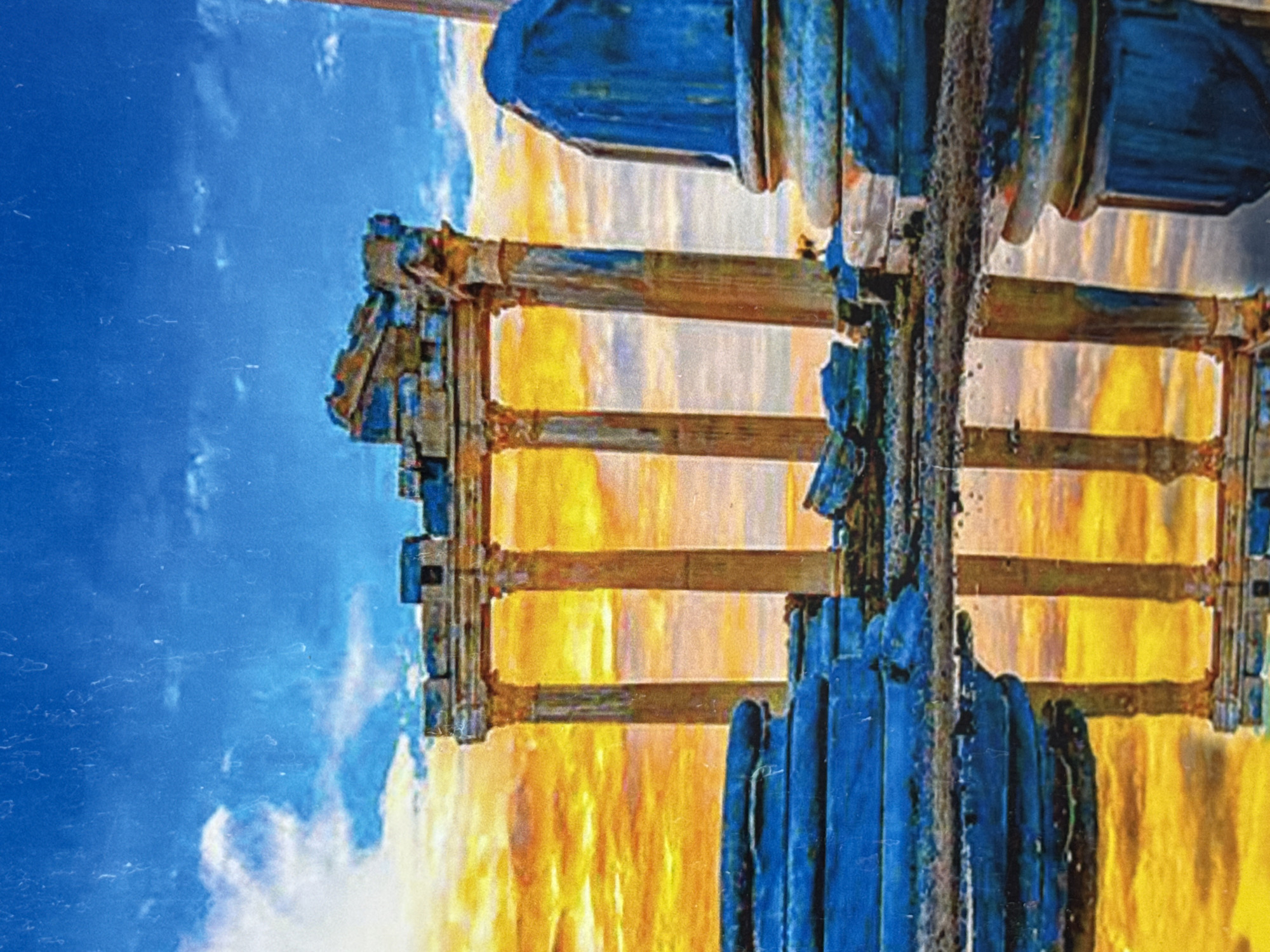 The image depicts a close-up, abstract-style view of what appears to be a section of the Alanya Teleferik (cable car) station in Alanya, Turkey. The scene shows a portion of the supporting structure, featuring wooden beams and possibly blue-painted metal components. A cloudy blue sky is visible in the background. The overall impression is of an aged or weathered structure, possibly captured with a lens that creates a softened or blurred effect.