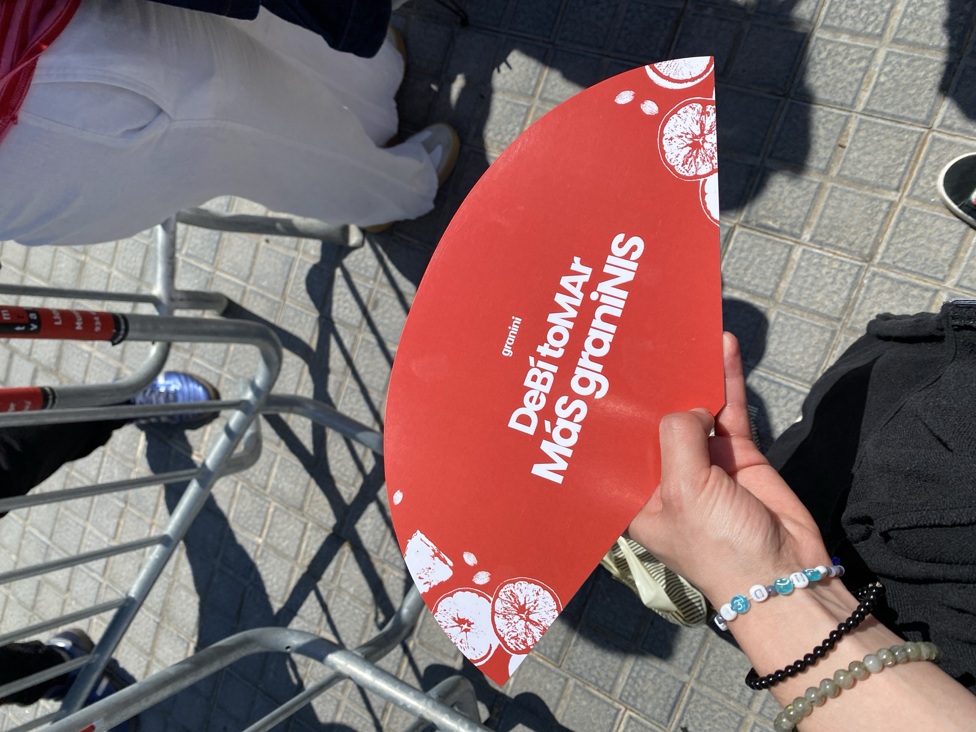A high-angle medium shot captures a hand holding a red, fan-shaped advertisement for a brand of juice. The ad features an image of cut citrus fruit and the phrase "DeBitoMAr Más graniNis". The hand is light-skinned and adorned with three bracelets. The background is composed of people, metal barriers, and stone-tiled ground with shadows, indicative of a sunny outdoor location. The presence of a metal barrier, casual attire of the surrounding people, and the tiled ground suggests a possible public space in an urban environment, possibly Barcelona, Spain.