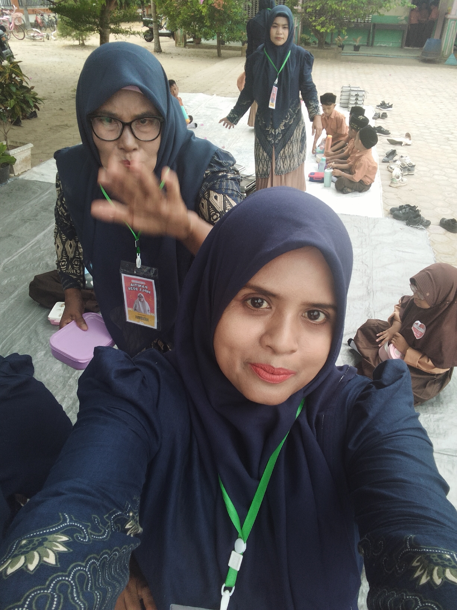 The image is a medium shot, taken from a low angle, of two Indonesian women wearing navy blue hijabs in what appears to be an outdoor school setting in Baratpayaitek, Indonesia. The woman in the foreground is positioned to the right of the frame, her face and chest filling the majority of the shot. She is looking directly at the camera, with bright red lipstick and dark eyes. She is wearing a navy blue hijab and a navy blue top with embroidered sleeves. A green lanyard with a white plastic clip is visible around her neck.
The second woman is in the background, slightly to the left and further away from the camera. She is also wearing a navy blue hijab and glasses. Her facial expression is unclear, but her right hand is raised, fingers spread, as if gesturing or waving. She is wearing a patterned top and a green lanyard with a badge. The badge is visible and appears to have a photo of a man and text in Indonesian.
Behind the women, several children in school uniforms are sitting on a white tarp on the ground. Some of the children are wearing orange shirts and dark pants, while one child wears a brown hijab and brown dress. There are various objects scattered around, including shoes, plastic containers, and what looks like stacks of books or binders. The environment suggests a schoolyard or common area, with sandy ground and some trees visible in the background. In the far background, there are buildings with green trim and bicycles. The overall scene appears to be a casual gathering or event at a school.