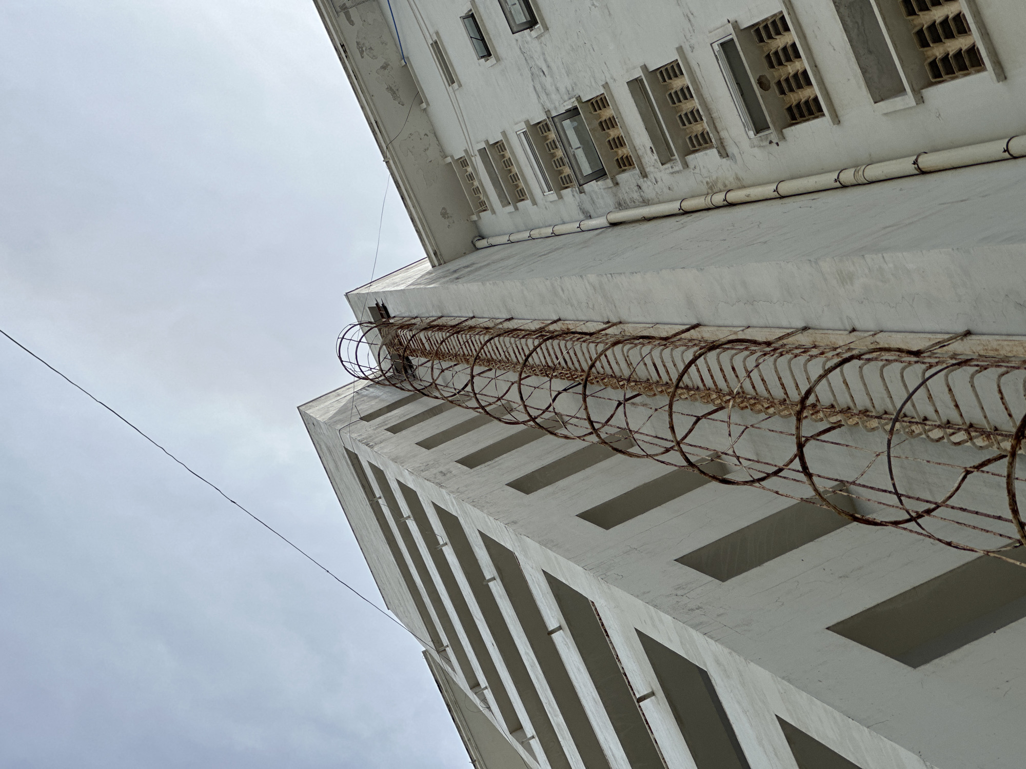 The image depicts the exterior of a weathered, multi-story building, seen from a low-angle perspective looking upwards. The building is predominantly white, with visible signs of age and possibly neglect, including some discoloration and hairline cracks on the facade.
On the left side of the building, a portion of what appears to be an external staircase or fire escape is visible. This structure is characterized by a rusty, spiral cage surrounding a ladder. The rust is a prominent feature, indicating significant exposure to the elements.
The building's architectural style suggests it could be an older residential or commercial structure. The windows are small and rectangular, some of them having louvers. There is a thin, dark cable stretched diagonally across the upper portion of the frame, disappearing behind the building. The sky above is overcast with a light, diffused gray tone.
The location provided is Nha Trang Ward, Vietnam. While the image itself doesn't contain explicit markers of this specific location, the architectural style and the general atmosphere are consistent with some urban environments in Vietnam. There are no people or animals visible in the image, and no discernible actions or interactions are taking place. The scene is static and focuses solely on the building's exterior and its immediate surroundings.