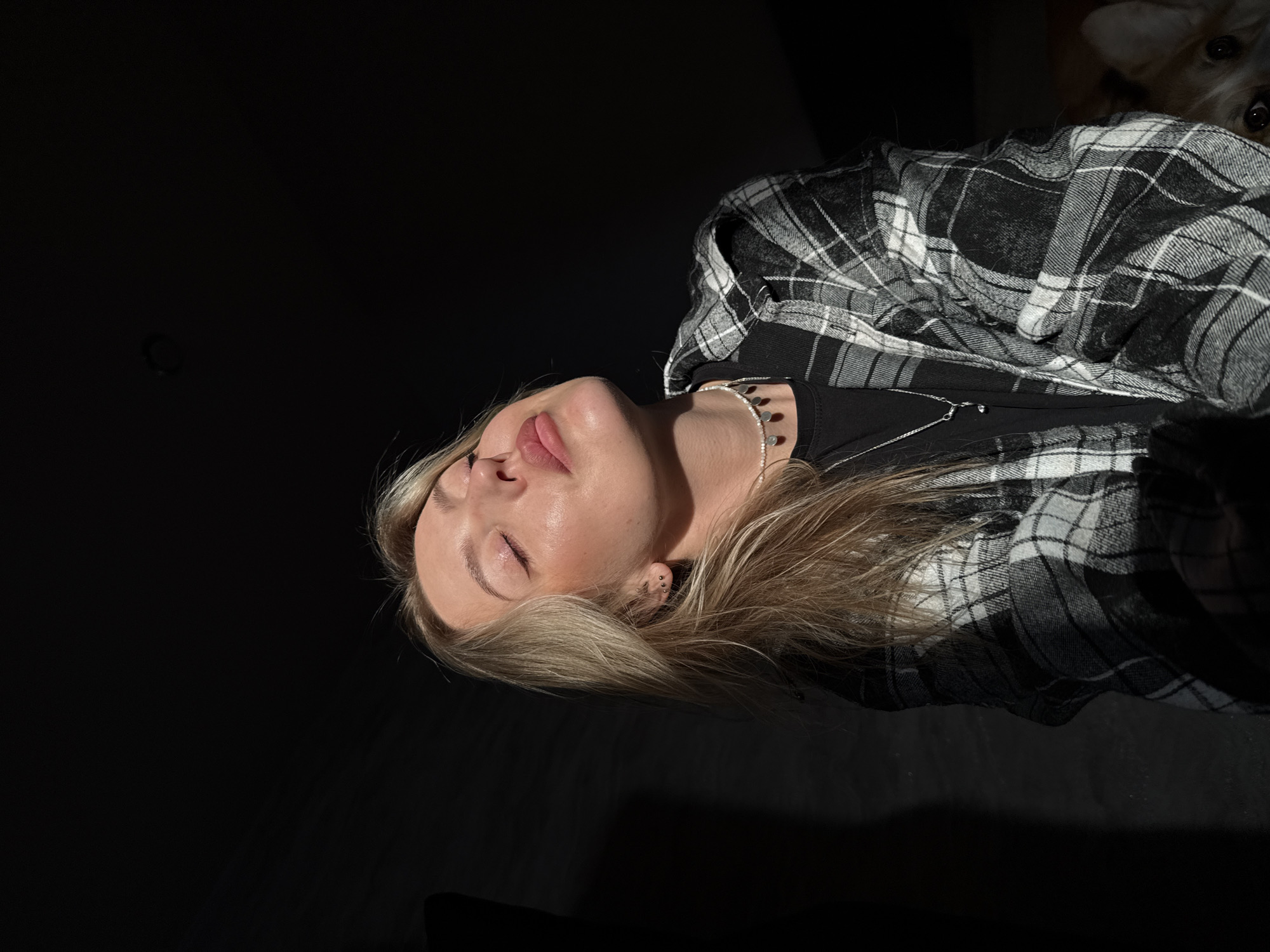 A woman with light blonde hair is lying down, eyes closed, in a dimly lit setting. She wears a black shirt underneath a black and white plaid flannel. She also wears two necklaces; one appears to be made of small beads and the other is a thin silver chain with a pendant. The scene is dominated by a large area of darkness, with the woman's face and upper torso illuminated by a direct light source. The location in the city of Krasnoyarsk, Russia is not visible.