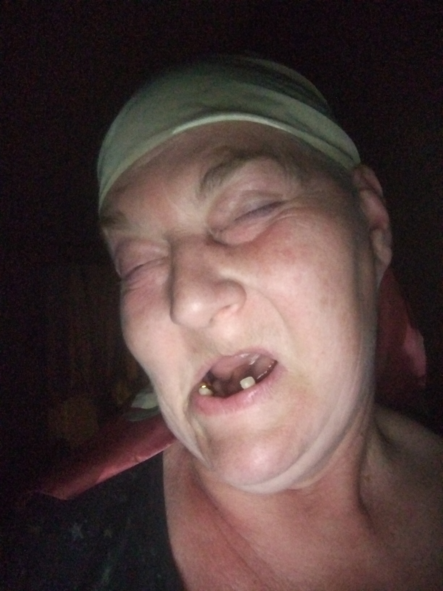 The image is a close-up, dimly lit, selfie-style photograph of a middle-aged Caucasian individual. The subject is wearing a light-colored bandana or headscarf and has their eyes closed, with their mouth open in a grimace. A gold-colored tooth is visible in the upper jaw. The background is dark and indistinct, suggesting an indoor environment with low lighting. There are no identifiable landmarks or context that would indicate a specific location in Horishni Plavni, Ukraine. The overall tone of the image is informal and candid.