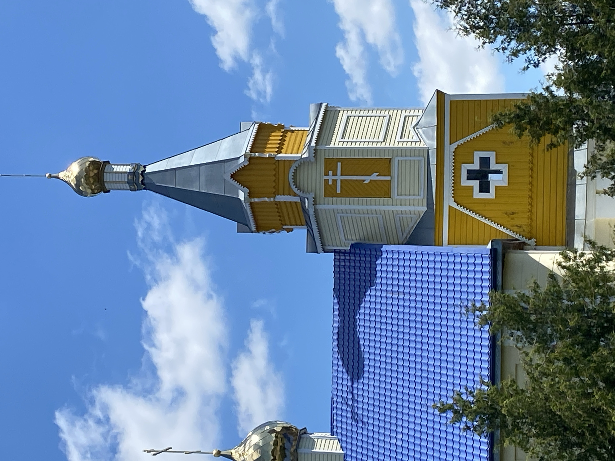 This is a medium-shot, eye-level, outdoor photograph of a church in Воўчынскі сельскі Савет, Belarus. The church has a prominent blue roof and a tall yellow tower topped with a golden dome and cross. The tower also features a cross symbol. The sky is blue with scattered white clouds. Evergreen trees are visible in the foreground. The lighting suggests it is daytime. There are no people visible in the image.