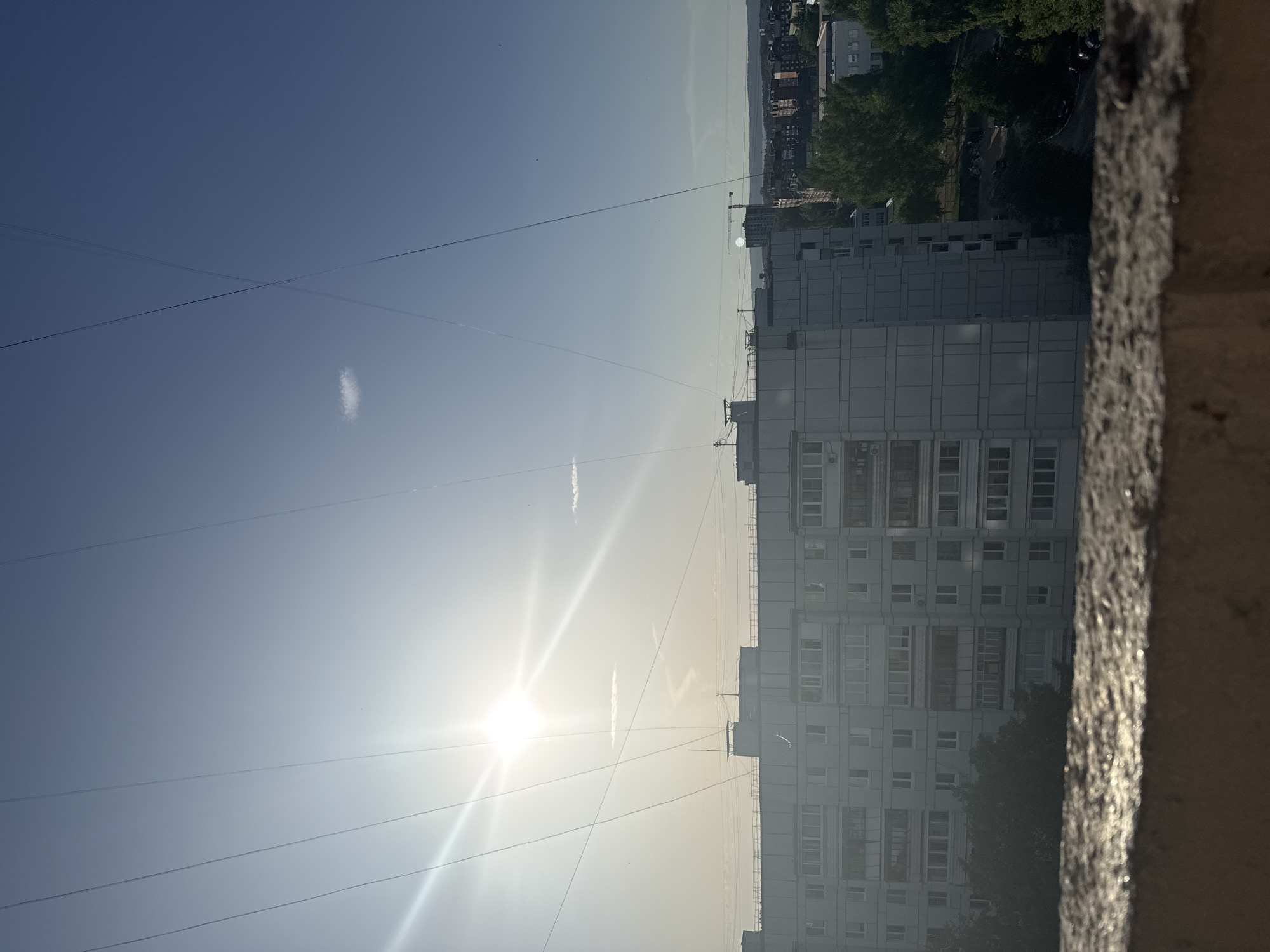 The image shows a view from a high vantage point, possibly a balcony, overlooking a cityscape in Chaikovsky, Russia. In the foreground, a rough concrete ledge is visible. Wires stretch across a bright, sunny sky, with the sun's glare prominent. A tall, residential building with multiple floors and windows dominates the middle ground. Trees and other buildings are visible further in the background. The image is taken from a tilted perspective, making it appear as if the scene is sideways. The overall impression is one of a hazy, bright day in an urban environment.