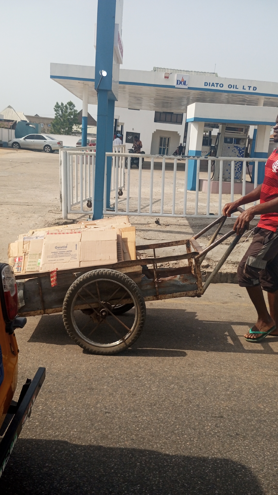 A man is pulling a two-wheeled cart loaded with cardboard boxes along a paved road in Lokoja, Nigeria. The boxes are labeled "Gestid" and bear the name "Ramsay World Limited." The man is wearing a red t-shirt, brown shorts, and green flip-flops. The cart is made of metal and has a rusty appearance. In the background is a gas station labeled "Diatto Oil LTD." There are other people and vehicles visible near the gas station.