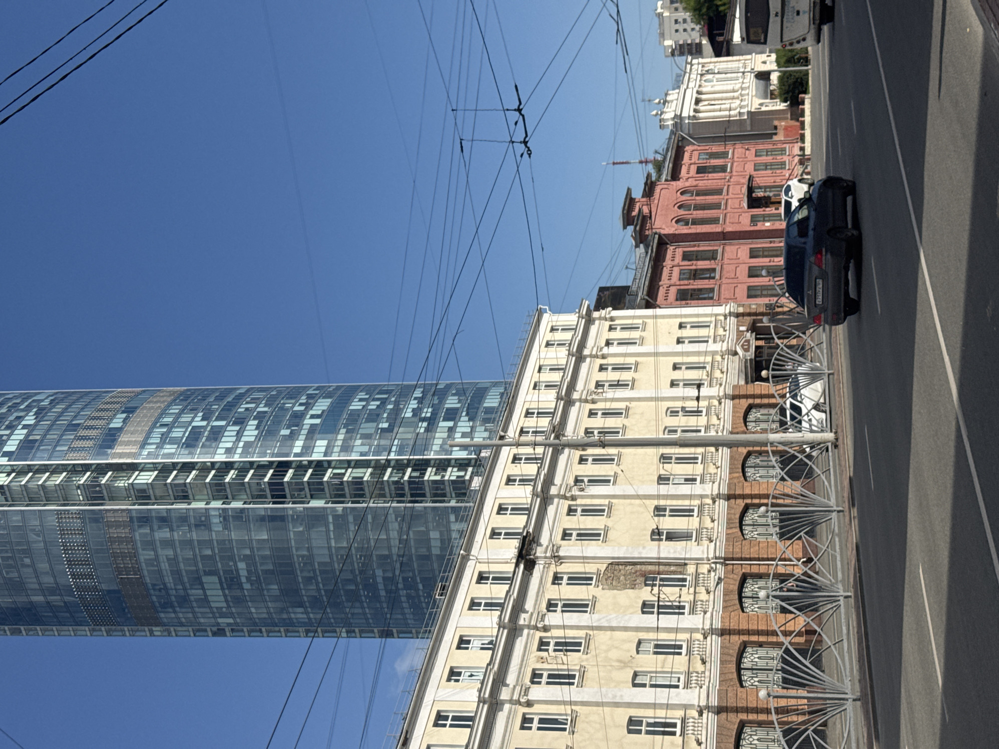 The image captures a street scene in Yekaterinburg, Russia, on a clear, sunny day. In the background, a modern, tall, glass-fronted skyscraper dominates the skyline. In the middle ground, a row of older, cream-colored buildings with ornate facades and numerous windows stretches along the street. To the right of these buildings, there are several other structures, including a vibrant pink building and a white building with classical architectural elements. Overhead, a complex network of electrical wires, likely for public transportation like trams or trolleybuses, crisscrosses the sky. In the foreground, a grey sedan is driving down the asphalt road. A white SUV is visible further down the road, and a white bus is on the right side of the frame, partially visible. The street is lined with a decorative metal fence. The overall impression is an urban environment where historical architecture coexists with contemporary development.