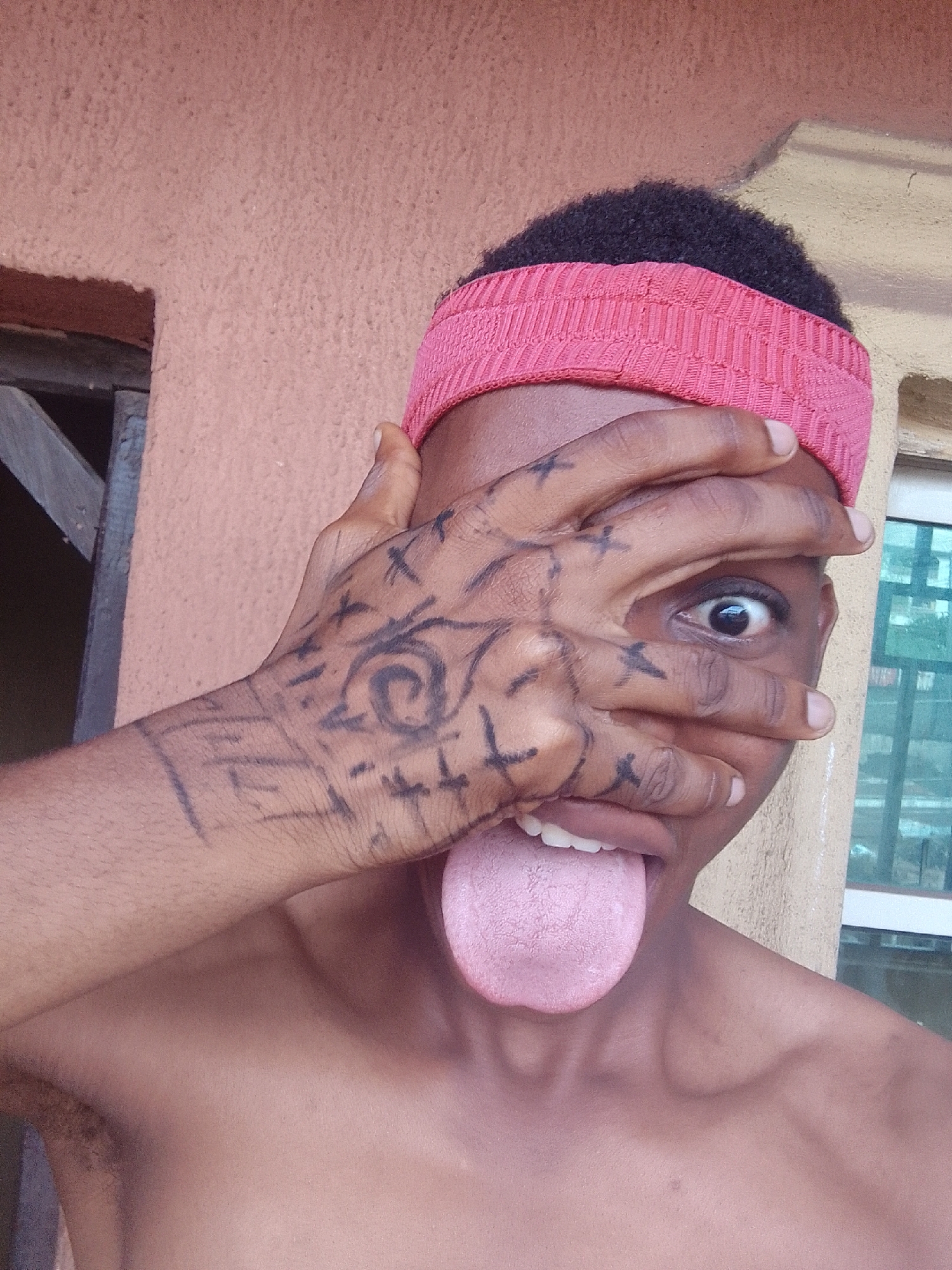 A person with dark skin, short black hair, and wearing a pink headband is visible from the chest up, facing the camera. They have drawn black markings on their right hand and arm, featuring swirling patterns and cross shapes. The person is sticking their tongue out and looking through their fingers with their right eye wide open. The background shows part of a building with a textured brown wall, a wooden structure, and a window with a white frame. The city and specific location are unknown.