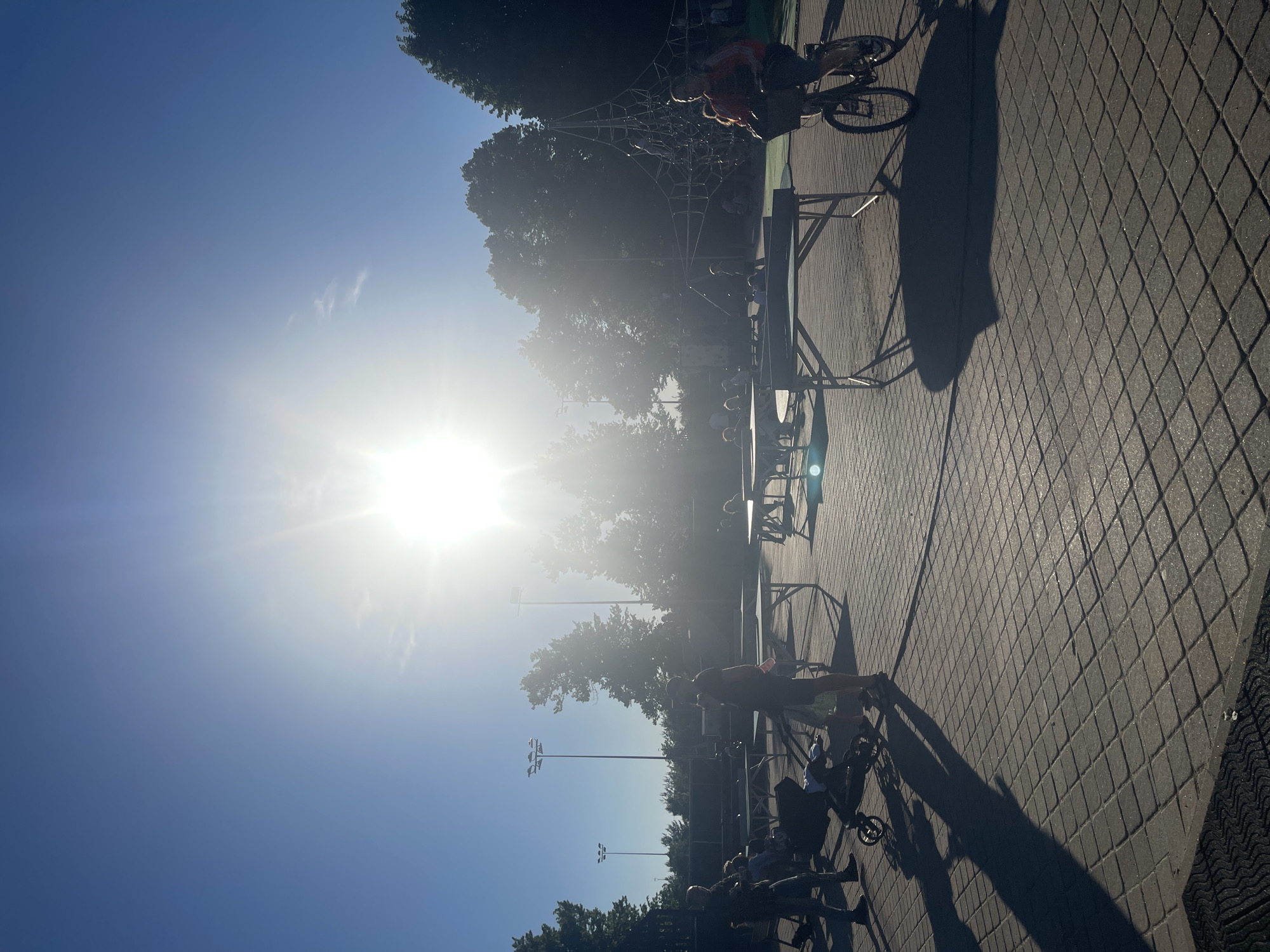 This is a low-angle, wide shot of an outdoor recreational area on a sunny day. The sun is intensely bright, positioned in the upper center of the frame, causing lens flare and washing out the sky. In the foreground, a paved area with a repeating square tile pattern is visible. To the left, a person stands near a stroller, casting a long shadow. In the middle ground, several folding tables are set up, some appearing to be ping pong tables. People are scattered throughout this area, some standing and others seated. To the right, a person rides a bicycle, also casting a long shadow. Behind the tables and people, there are trees and what appears to be a playground structure with a net. Tall lampposts are also present in the background. The overall impression is of a public space being used for leisure activities.