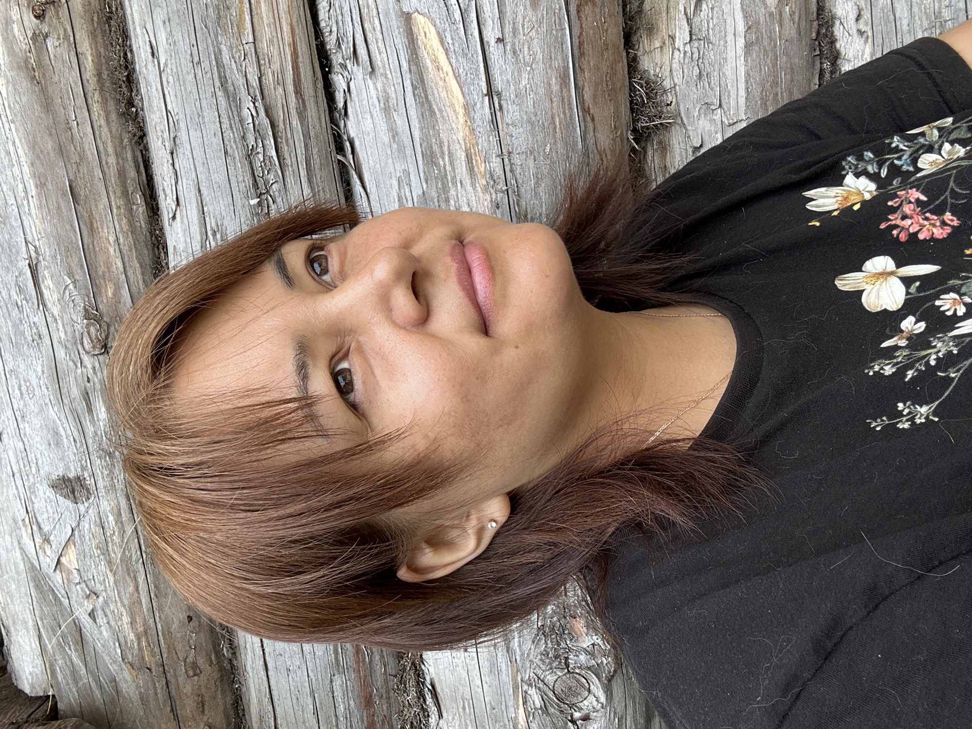 A medium shot shows a woman from the chest up, looking up and to the right, with a weathered wooden wall as the background. She has brown hair with reddish highlights, pulled back from her face, and fair skin with a light smattering of freckles. She is wearing a black t-shirt with a delicate floral pattern of white and pink flowers. A thin gold chain is visible around her neck. Her left ear, adorned with a small, clear stud earring, is visible. The wooden wall behind her is made of rough, horizontal logs with visible bark and cracks. The lighting is natural and appears to be from an overhead source.