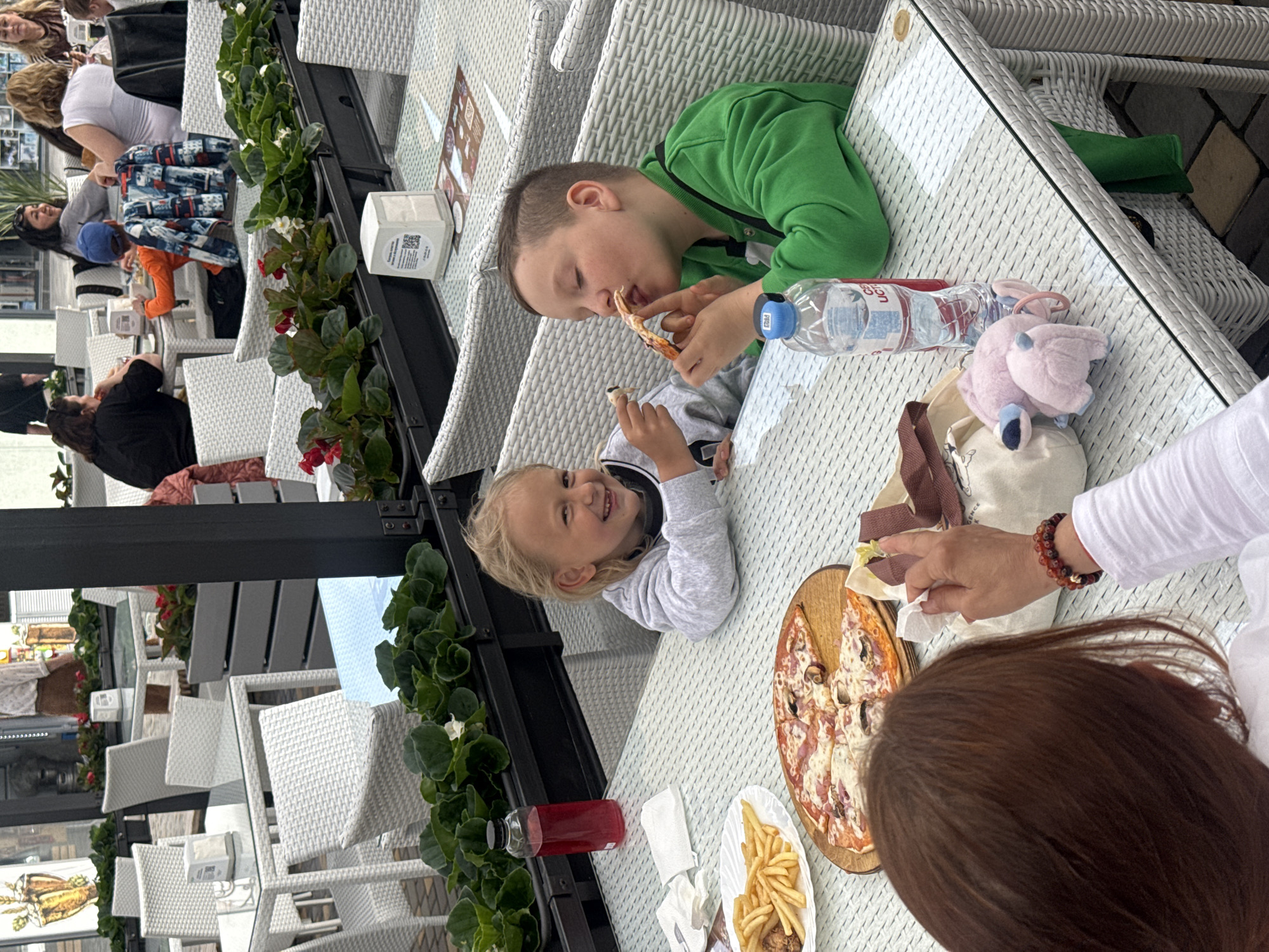 Here is a detailed description of the image:
**Scene:** The image shows an outdoor dining scene, likely at a restaurant in an unspecified city.
**People:** Two children, a boy and a girl, are the central subjects. The boy wears a green shirt and is eating a piece of pizza. The girl has blonde hair and is smiling at the camera while holding a piece of pizza. Part of an adult woman is visible in the lower right, with brown hair and a white shirt. Other people are visible seated at tables in the background.
**Objects:** A table is the central feature, covered with various food items, including a pizza on a wooden platter, a plate of french fries, and two beverage bottles. There's also a pink plush toy and a handbag on the table. The table and chairs are made of woven white material. Decorative flower pots with green plants and red flowers are arranged along the edge of the table, likely part of the restaurant's decor.
**Actions:** The boy and girl are eating pizza. The adult woman is using a napkin.
**Interactions:** The boy and girl appear to be enjoying their meal.
**Environment:** The restaurant appears to have an outdoor seating area, possibly along a street or promenade. The lighting suggests it is daytime. The furniture design gives the impression of a modern, casual dining environment.