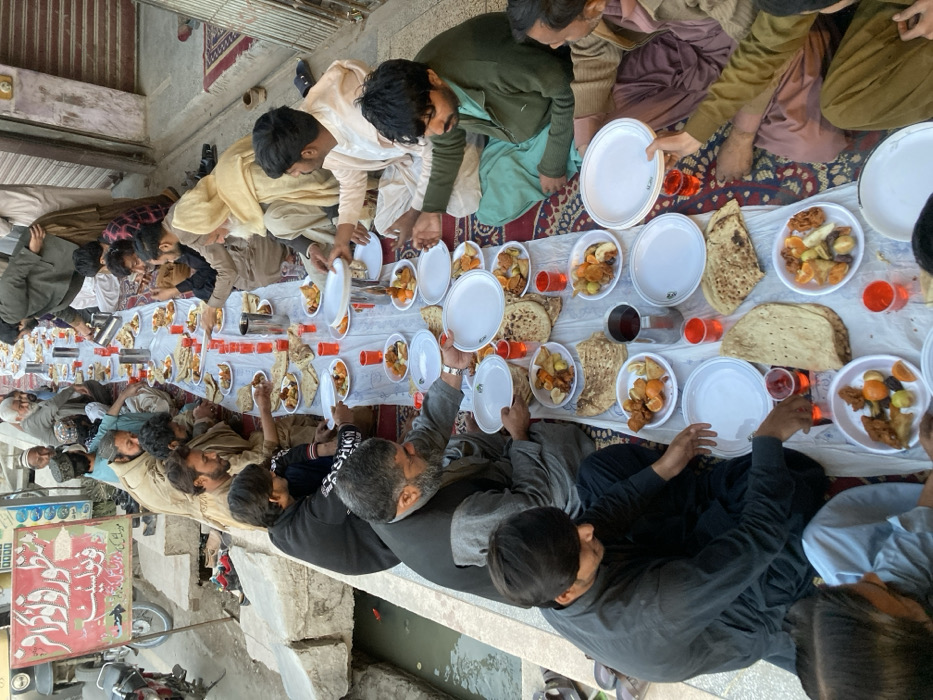 Approximately 20-25 adult men are engaged in an outdoor communal meal in a street or alleyway within Quetta City Tehsil, Pakistan. The men are seated on patterned carpets laid over the pavement, along both sides of a long, white disposable plastic sheet that serves as a communal table.
The table is densely covered with white disposable plates, each containing various food items including flatbread, fried snacks, sliced mandarins, dates, and other small items. Red plastic cups filled with a red beverage and several silver metallic jugs are interspersed among the plates. Several men are actively eating, while others are holding or passing stacks of empty white disposable plates. The men are primarily dressed in traditional South Asian attire, including shalwar kameez and some wearing waistcoats.
To the left of the seated group, an open concrete drainage ditch containing dark water and some debris runs parallel to the meal area. Beyond this ditch, a parked motorcycle is visible, and a white sign with red and green Urdu script is affixed to a pole or building. To the right, a building with a closed corrugated metal shutter, indicative of a storefront, is present. The scene is illuminated by natural daylight.