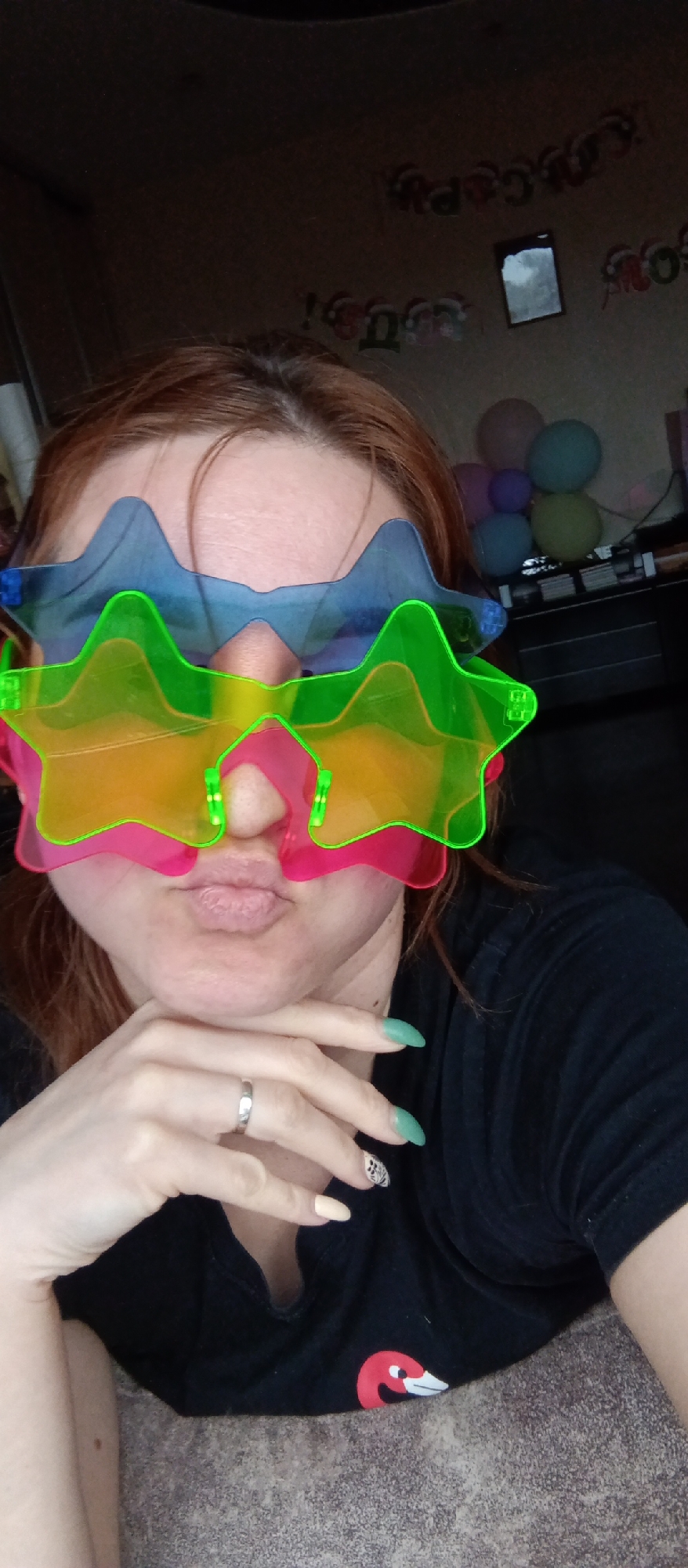 A person is indoors, posing for a selfie. They are wearing two pairs of star-shaped novelty sunglasses, one blue and one green. The person is wearing a black T-shirt with a red and white graphic on it. Their fingernails are painted in various shades of green and white. They are wearing a plain silver ring. In the background, there are some balloons and a "Happy Birthday" banner.