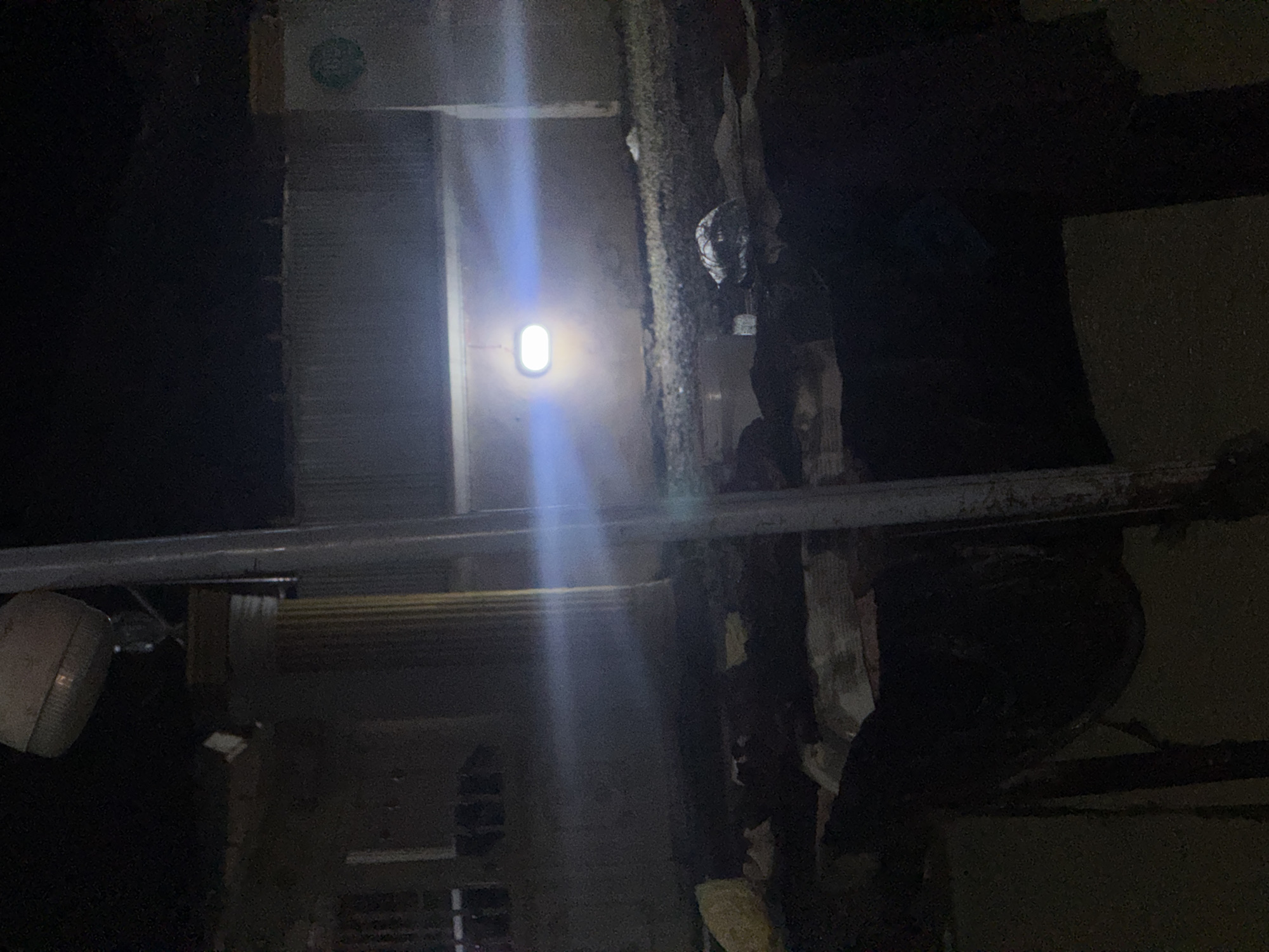 In the city of Abuja, Nigeria, at night, a scene of apparent destruction and abandonment is depicted. The foreground is cluttered with debris, including what appear to be large bundles wrapped in dark material and scattered papers or fabric. A wooden structure, possibly a makeshift table or stand, is visible amongst the debris. In the mid-ground, a building with a large, white, oval-shaped light illuminates the scene, casting a bright glow. The building itself appears to be constructed of concrete and features a fence or wall. A tall, grey pole stands prominently in the mid-ground, bisecting the image vertically. In the background, a large, imposing structure, possibly a pillar or column, is visible to the left, topped by a light fixture. The overall impression is one of disarray and neglect, with the artificial lighting creating stark contrasts and long shadows.