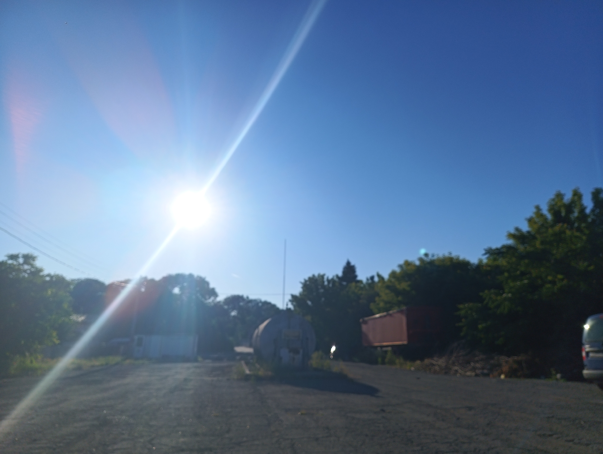 The image captures a sunlit outdoor scene, likely in an industrial or undeveloped area, possibly on the outskirts of Karnobat, Bulgaria, given the provided context. A bright sun is visible in the upper left quadrant, casting strong lens flares across the sky and into the foreground. The sky is a clear, bright blue, with no clouds.
In the foreground, a rough, dark-colored surface, appearing to be gravel or dirt, dominates the lower half of the image. This surface suggests a road or an open lot. To the left, a thin pole with power lines stretches across the scene.
Several structures are present in the middle ground. A white, rectangular building with a dark door is situated on the left side, partially obscured by trees. To the right of this building, a large, cylindrical, tank-like object with a distressed surface and possibly some markings is positioned on the ground. Further to the right, a reddish-brown, rectangular shipping container is visible. Behind these structures, a dense line of green trees forms a backdrop, with some taller trees and foliage extending into the upper right portion of the image.
On the far right edge of the frame, a dark-colored vehicle, appearing to be an SUV or van, is partially visible, facing away from the viewer. Its rear section, including a taillight, is discernible. The overall impression is of a somewhat desolate, open area bathed in bright daylight.