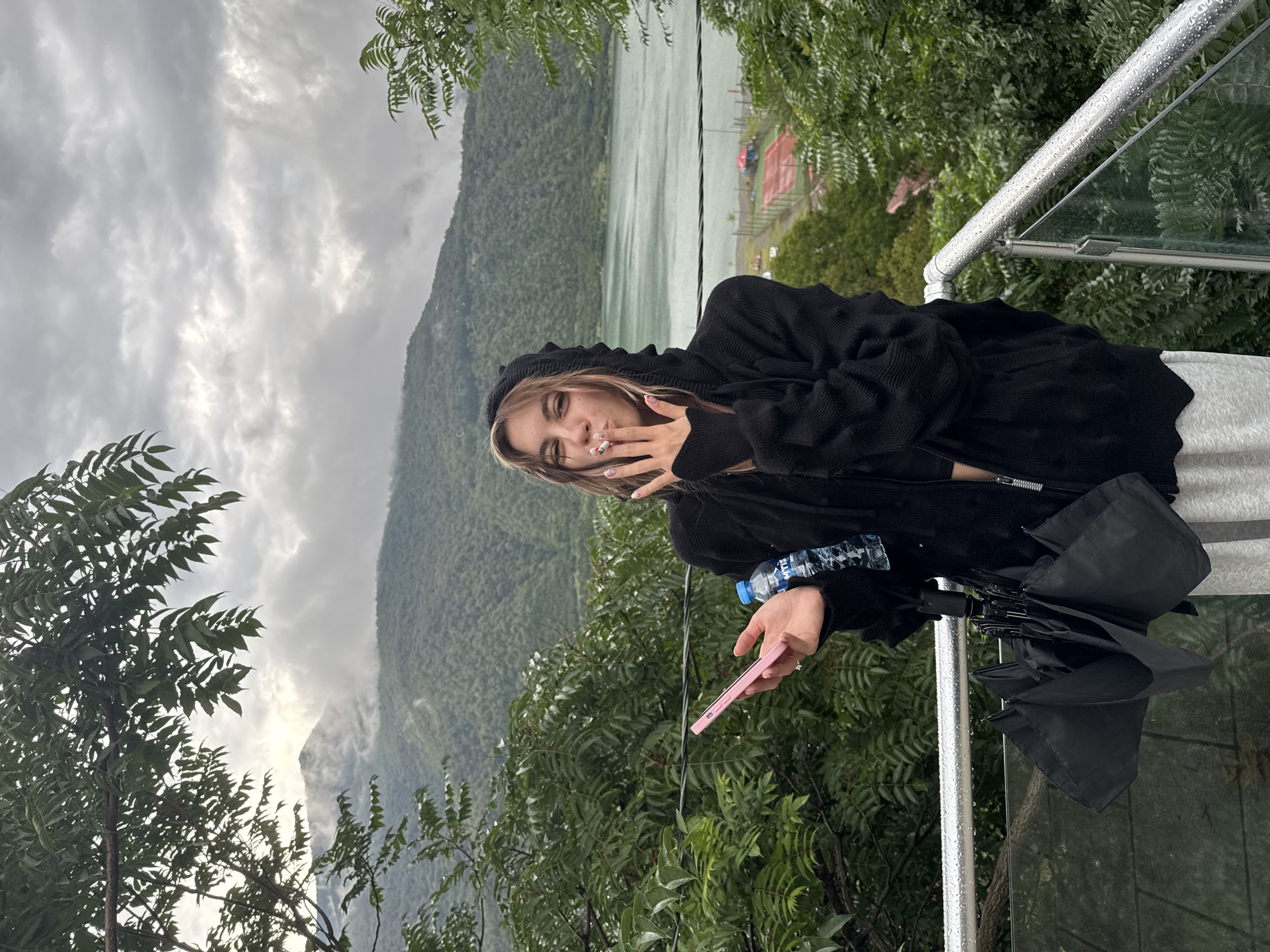 A young woman stands on a balcony overlooking a lake and mountains. She is wearing a black hooded sweater and gray sweatpants, and she is smoking a cigarette. In her left hand, she holds a pink smartphone and a bottle of water. A black umbrella is tucked under her left arm. The background shows a cloudy sky, green mountains, and a body of water. In the distance, there is a sports complex with tennis courts. The scene appears to be in Shalikiantkari, Georgia, as indicated by the reference to the location.