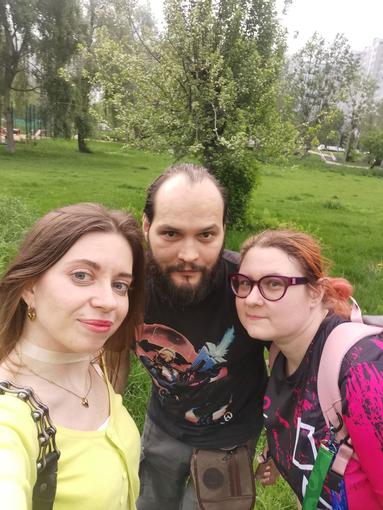 Three individuals are captured in a selfie outdoors. On the left, a woman with brown hair is wearing a yellow cardigan, a choker, and a necklace with a heart pendant. In the center, a man with a beard and dark hair is wearing a black graphic tee. On the right, a woman with red hair and glasses is wearing a black and pink shirt and a pink backpack. The background reveals a grassy park with trees. In the distance, buildings are visible. The location is in the city of Unknown, Unknown.