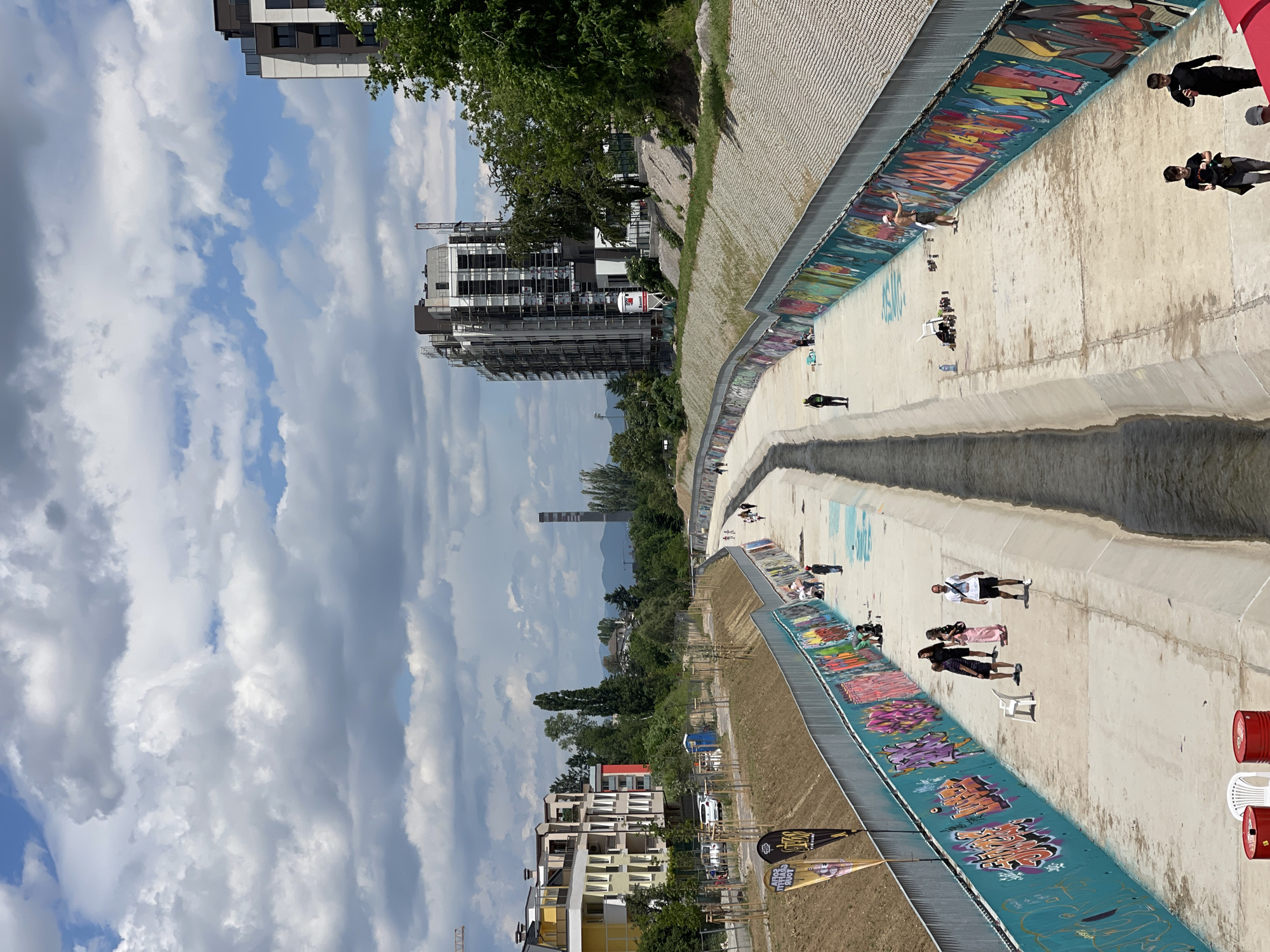 The image depicts a wide, concrete riverbed in Sofia, Bulgaria, transformed into a public space. The channel is lined with colorful graffiti. Numerous people are seen walking, relaxing, and sunbathing along the concrete surface. There is an adjacent landscaped area with trees and grass alongside the riverbed. In the background, buildings and a smokestack are visible against a partly cloudy sky. Red barrels and white chairs indicate temporary seating areas.