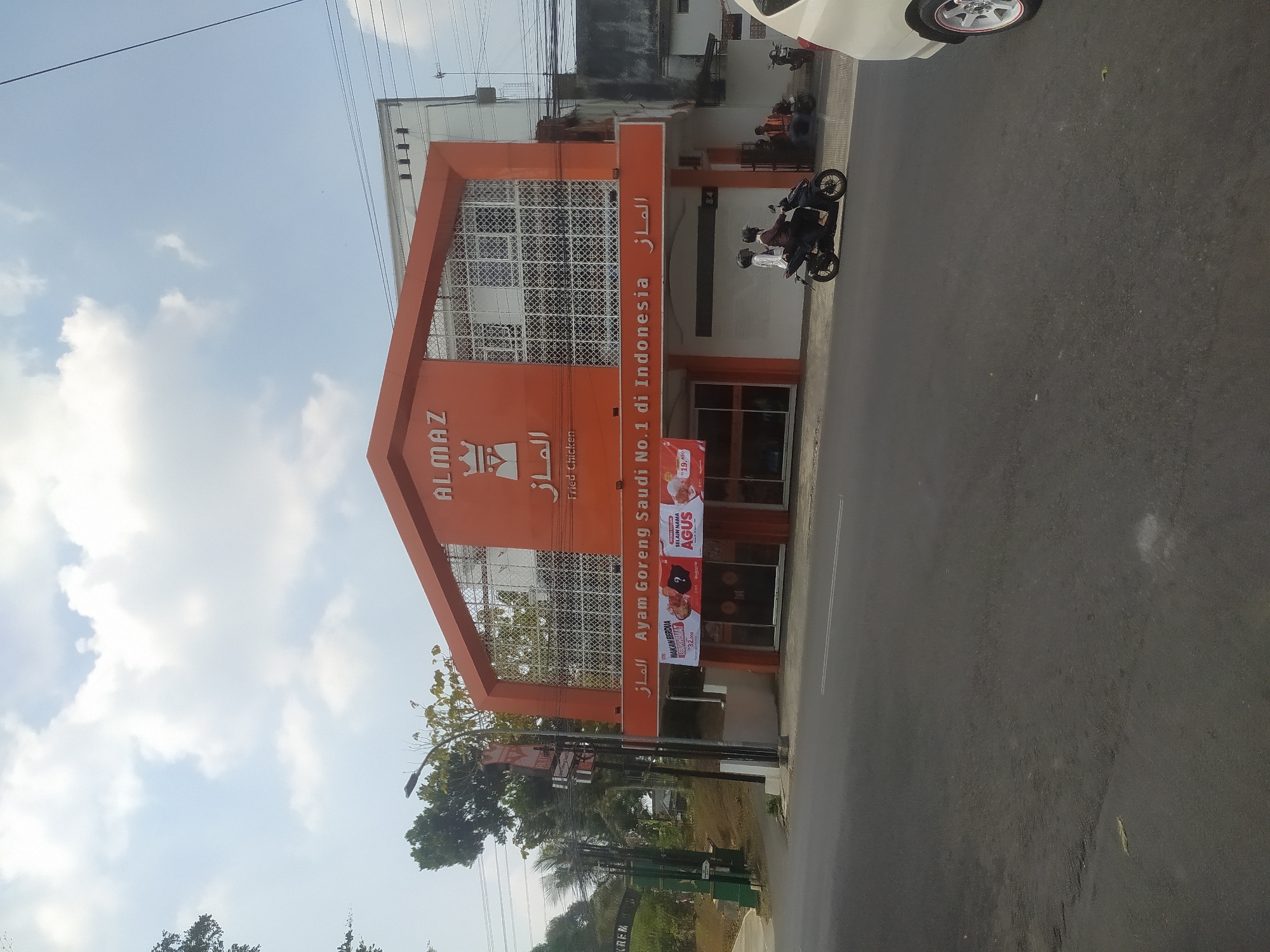 The image shows the exterior of an Almaz Fried Chicken restaurant, which claims to be the number one fried chicken in Indonesia. The building is painted orange and white, with Arabic script above the entrance. There are several banners outside advertising promotions, including one for "Makan Berdua Lebih Hemat" (Eating Together is More Economical) and another mentioning "AGUS". Two people are seen riding a motorcycle in front of the restaurant, and another person is sitting on a motorcycle to the right. A white car is partially visible in the foreground. The restaurant is located on a street with a paved road and sidewalks, and there are trees and power lines in the background. The sky is partly cloudy. The building's signage indicates it is located in Salatiga, Indonesia.