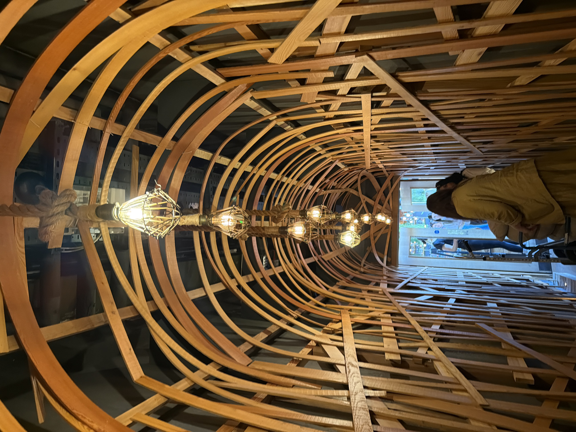 The image captures an intricately designed indoor space, likely a commercial establishment, characterized by an elaborate wooden tunnel structure. The perspective looks down the length of this structure, which is formed from numerous light-colored wooden planks and strips. These wooden elements are interwoven, with some forming graceful arches that create the tunnel's curved shape, while others run straight, creating a complex, skeletal pattern reminiscent of a boat's hull or rib cage.
Running through the center of this wooden tunnel is a string of decorative lights. Each light fixture consists of an exposed, glowing Edison-style bulb encased within a metal cage, and wrapped in thick rope, adding to a rustic or industrial-chic aesthetic. These lights emit a warm, orange glow, illuminating the wooden structure and suggesting an intimate, atmospheric ambiance, likely during the evening or in a dimly lit interior.
At the far end of the tunnel, two individuals are visible. They are seen from behind, facing away from the camera, and appear to be standing or walking towards a brighter area. One person wears a light brown or tan jacket. Beyond them, through an opening, another section of the establishment is glimpsed, showing more people, what appears to be a counter or display area, and a rectangular feature that could be a window or a framed picture depicting green foliage. The setting suggests a unique restaurant, bar, or gallery in London, UK, designed to create a distinctive visual experience for its patrons. No visible text is present in the image.