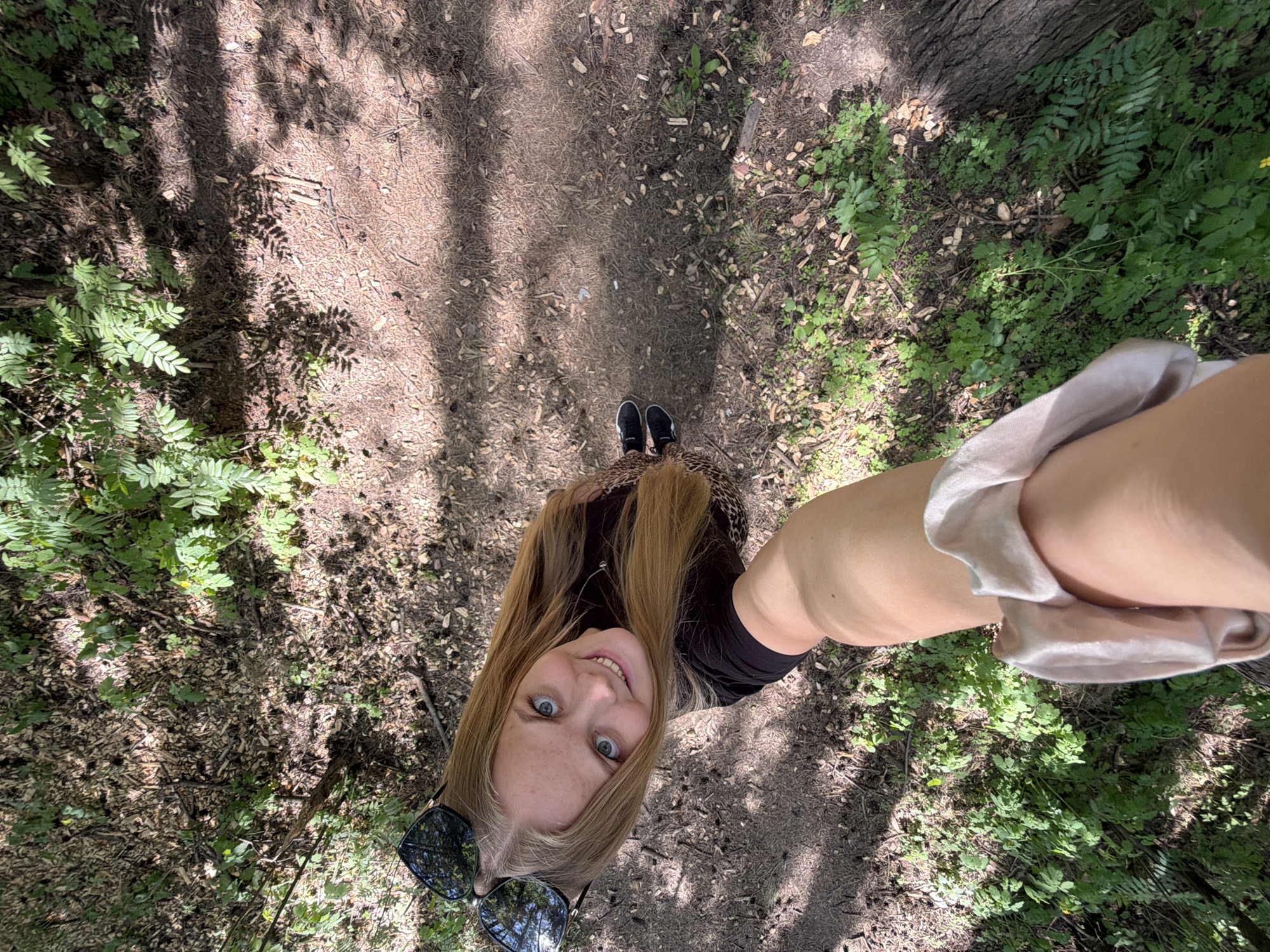 A young woman with long blonde hair is taking a selfie in a wooded area in Izhevsk, Russia. She is wearing a black top, a leopard print skirt, and black sneakers. Sunglasses are perched on her head. She is standing on a dirt path surrounded by trees and greenery. The photo is taken from a high angle, looking down at the woman.