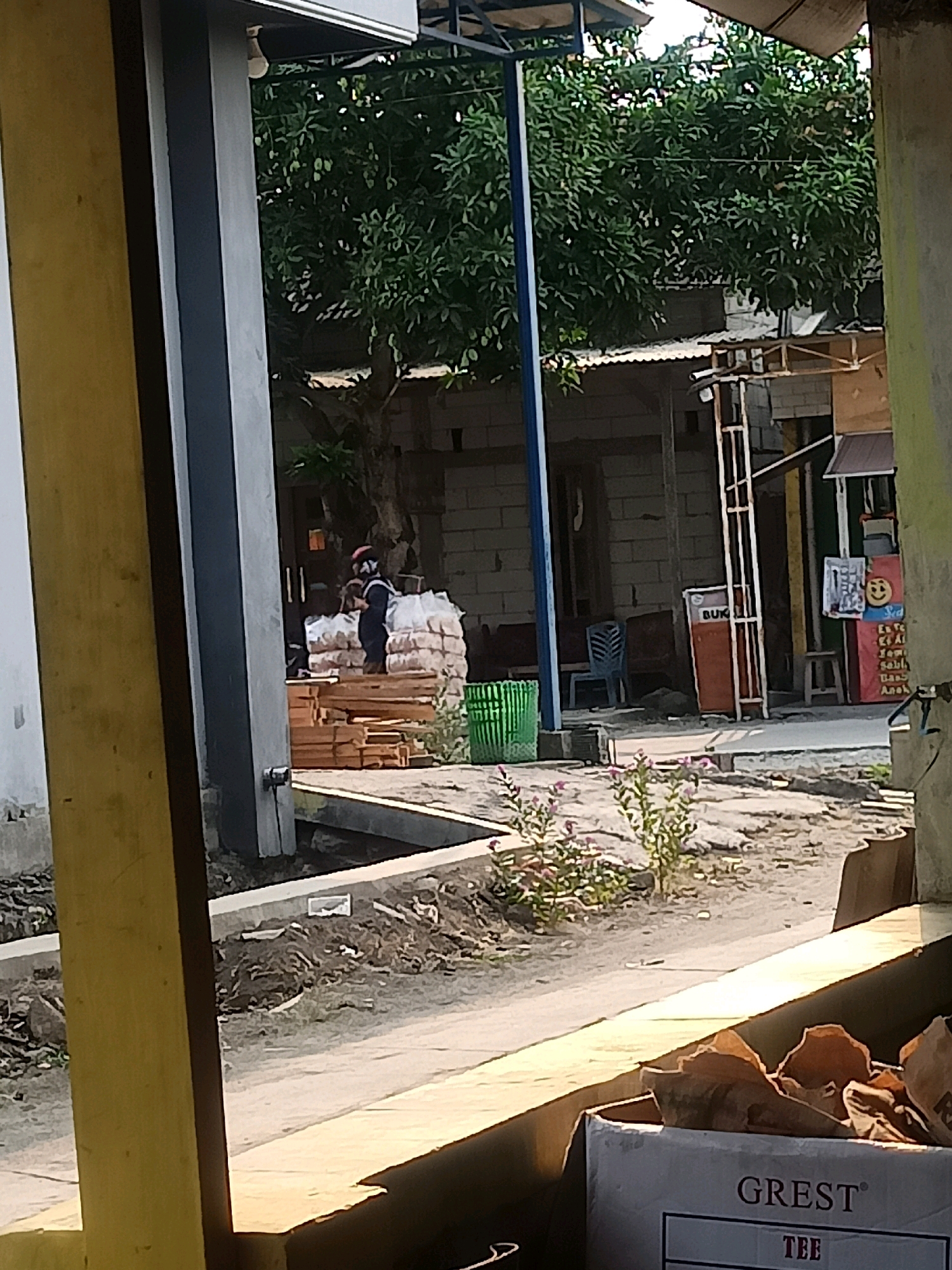The image depicts an outdoor scene in what appears to be a semi-built area, possibly a construction site or a marketplace, in Karangtalun, Indonesia. In the foreground, a bright yellow pillar is partially visible on the left, and a portion of a cardboard box with the text "GREST TEE" is in the lower right corner.
Behind these elements, a person wearing dark clothing and a red head covering is standing next to a large stack of white sacks, likely cement or building material, and a pile of wooden planks. The person is partially obscured by the sacks and planks.
Further in the background, there is a building with exposed brick walls and a corrugated metal roof. A blue chair and a green plastic basket are visible near the building. Signs are mounted on a structure to the right of the building, one of which features a smiling face emoji. Trees with dense foliage are present behind the structures, suggesting a tropical environment. The ground is covered with dirt and debris. Sunlight casts shadows on the ground, indicating it is daytime.