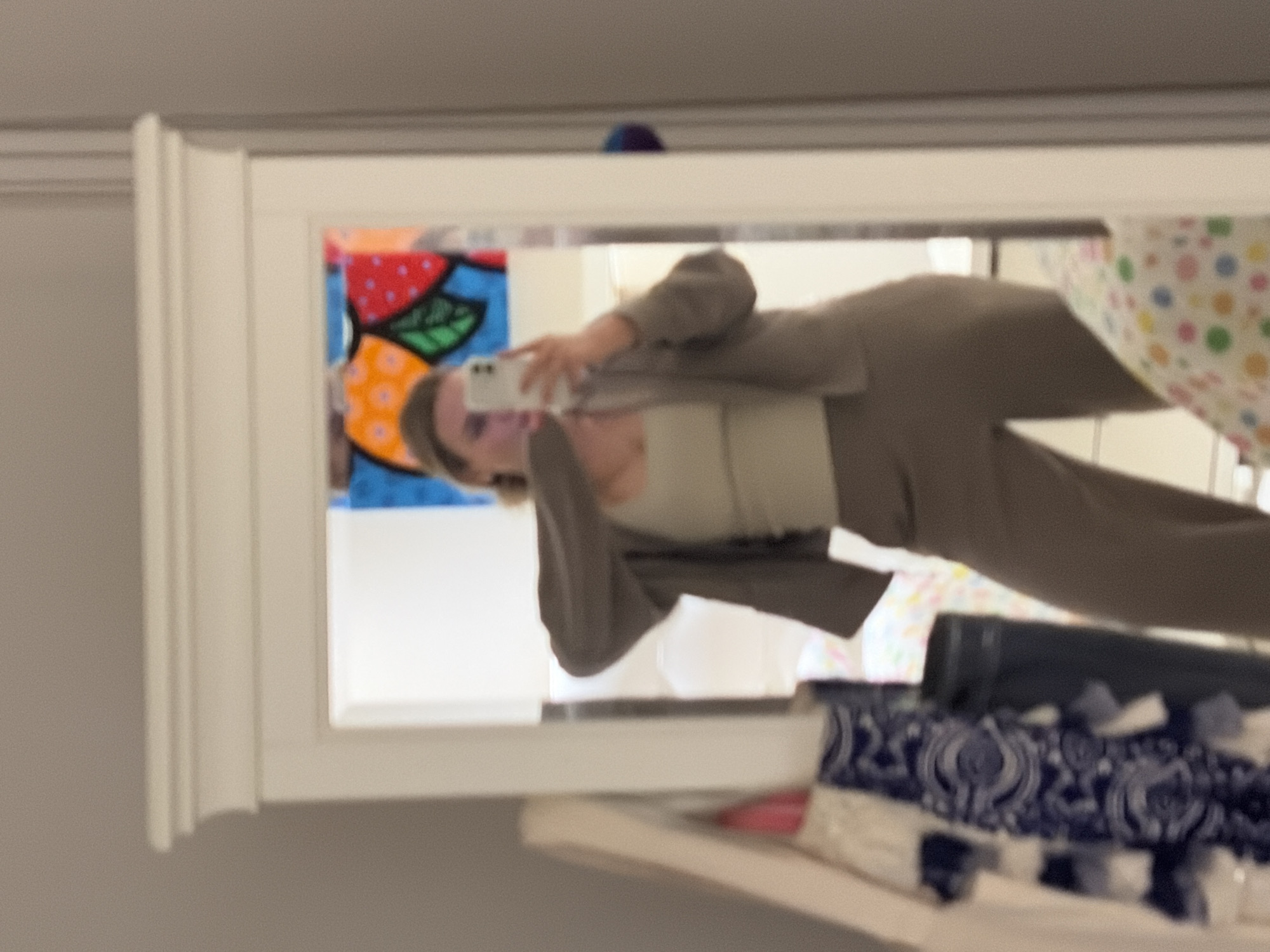 A person is reflected in a mirror while holding a smartphone and taking a selfie. The person is wearing a light-colored top and a brown blazer and pants. The mirror has a white frame and is positioned on a shelf with assorted items. In the background of the reflection is an abstract painting with vibrant colors, as well as what appear to be other objects and furnishings. The location is not possible to determine based on image content.