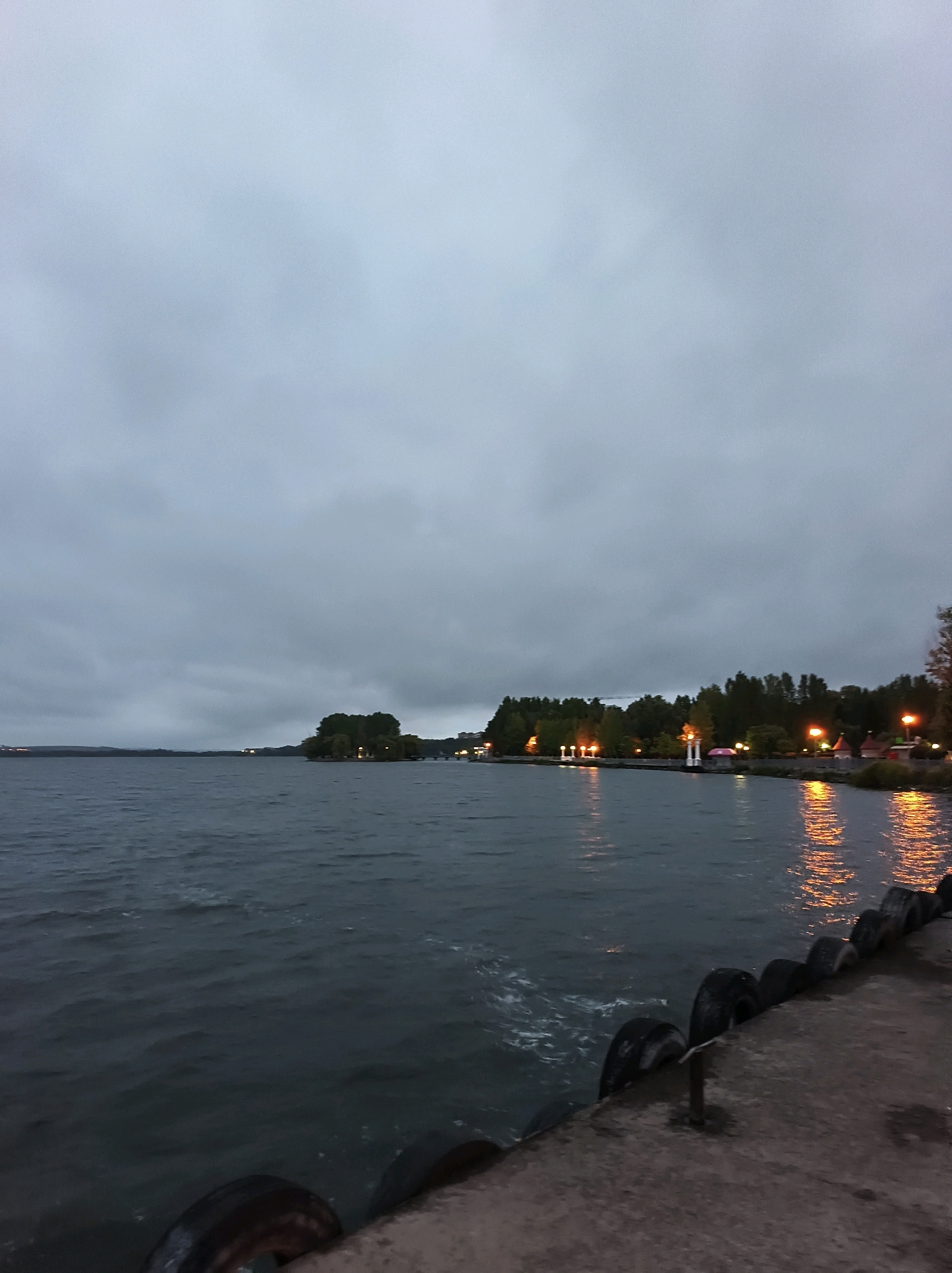 The image depicts a gloomy, overcast evening scene of the Ternopil pond in Ternopil, Ukraine. A concrete embankment with tires lining its edge occupies the foreground, leading the eye towards the choppy water. Across the pond, a distant treeline is illuminated by scattered artificial lights, suggesting a populated area or park. The sky is a uniform gray, with the water reflecting the limited light from the shore. No people or active events are visible.