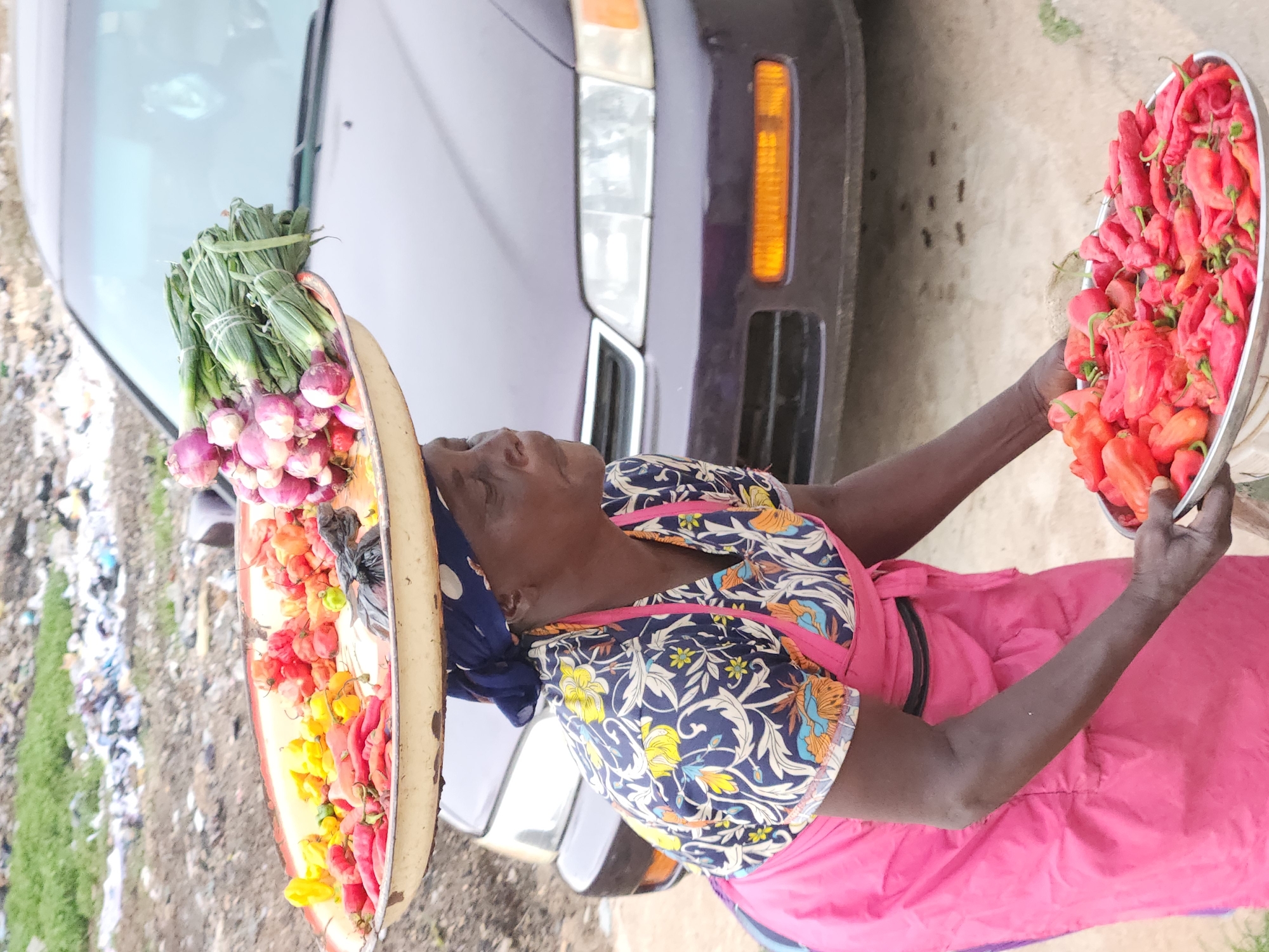 An elderly dark-skinned woman is standing outdoors next to a purple car. She wears a blue floral patterned blouse, a pink apron, and a blue headwrap. On her head, she balances a large, round tray laden with green onions and red onions, as well as red, orange, and yellow chili peppers. She also holds another tray filled with only red chili peppers. There is trash on the ground. The location is likely the city of Jos, Nigeria.