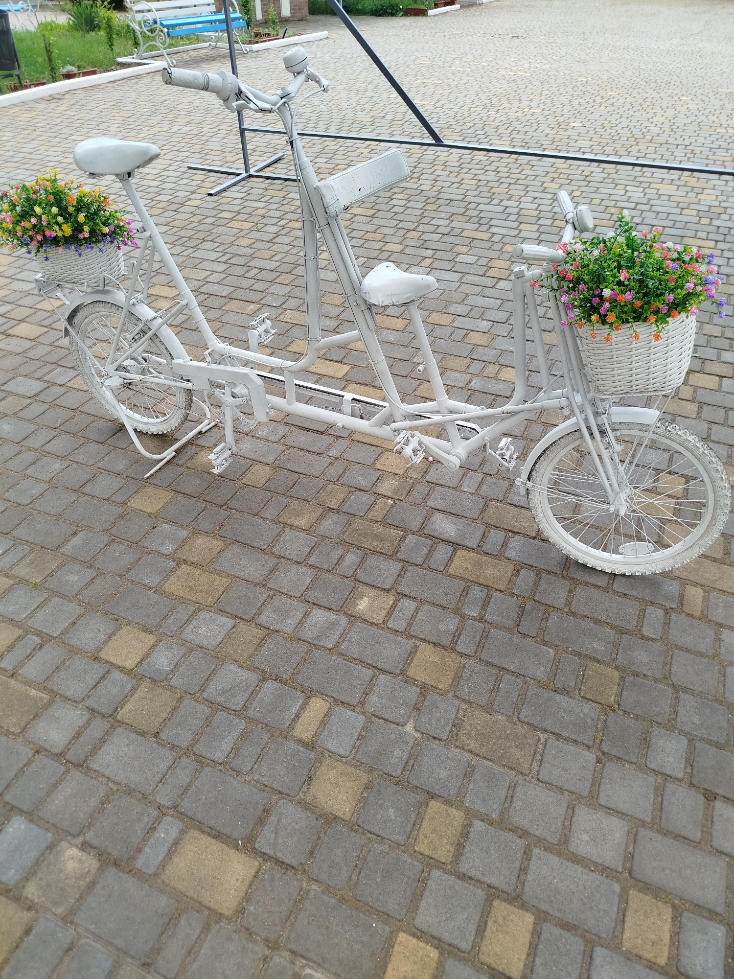 A white tandem bicycle, likely a decorative item, is positioned on a paved area. The bicycle features two seats and two sets of pedals, suggesting it's designed for two riders. White wicker baskets are attached to the front and rear of the bicycle, each filled with an assortment of colorful artificial flowers. The bicycle itself appears to be painted entirely white, including its tires. The ground beneath it is paved with rectangular cobblestones in varying shades of grey and tan. In the background, a white and blue bench is visible, along with some greenery and a dark metal structure. The overall impression is of an outdoor decorative setup, possibly in a park, garden, or public square.