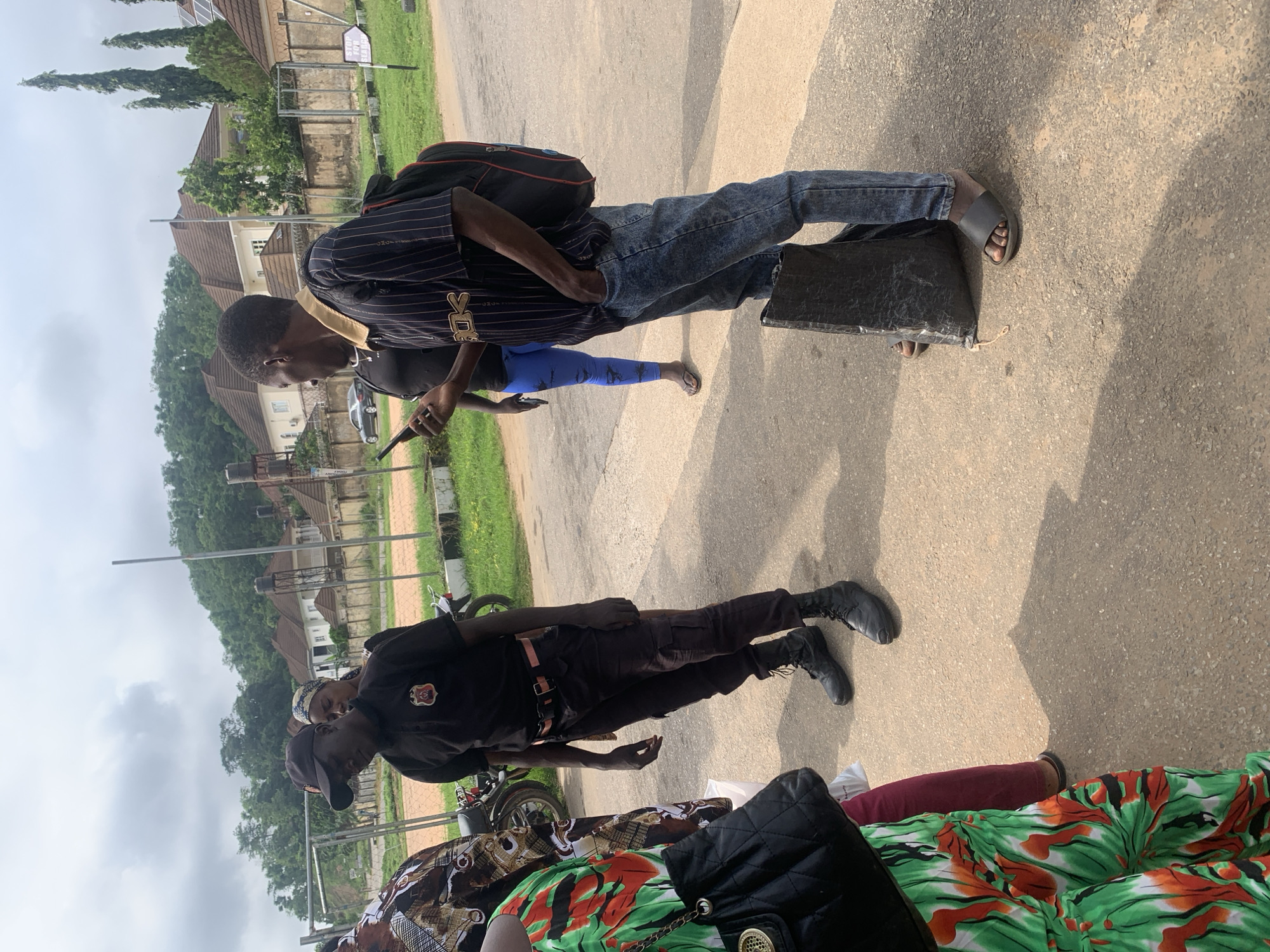 An outdoor scene, likely in Abuja, Nigeria, shows several individuals on a paved surface with grassy areas and buildings in the background. In the foreground, a person in a black uniform, including a cap, appears to be a security guard. They are wearing black tactical pants and black boots, with a belt and what may be a sidearm holster. To their left, another individual is partially visible, wearing a colorful dress with green, red, and black patterns. Behind the security guard, a woman with a patterned headscarf is seen. To the right, a man in a striped t-shirt and blue jeans is holding a mobile phone and has a backpack slung over one shoulder. Another person in blue leggings and a dark top is standing behind him. A motorcycle is parked to the left, and a sign with "STOP FOR CHILDREN" is visible in the background on the right. The environment suggests a residential or institutional area with some vegetation and fencing.