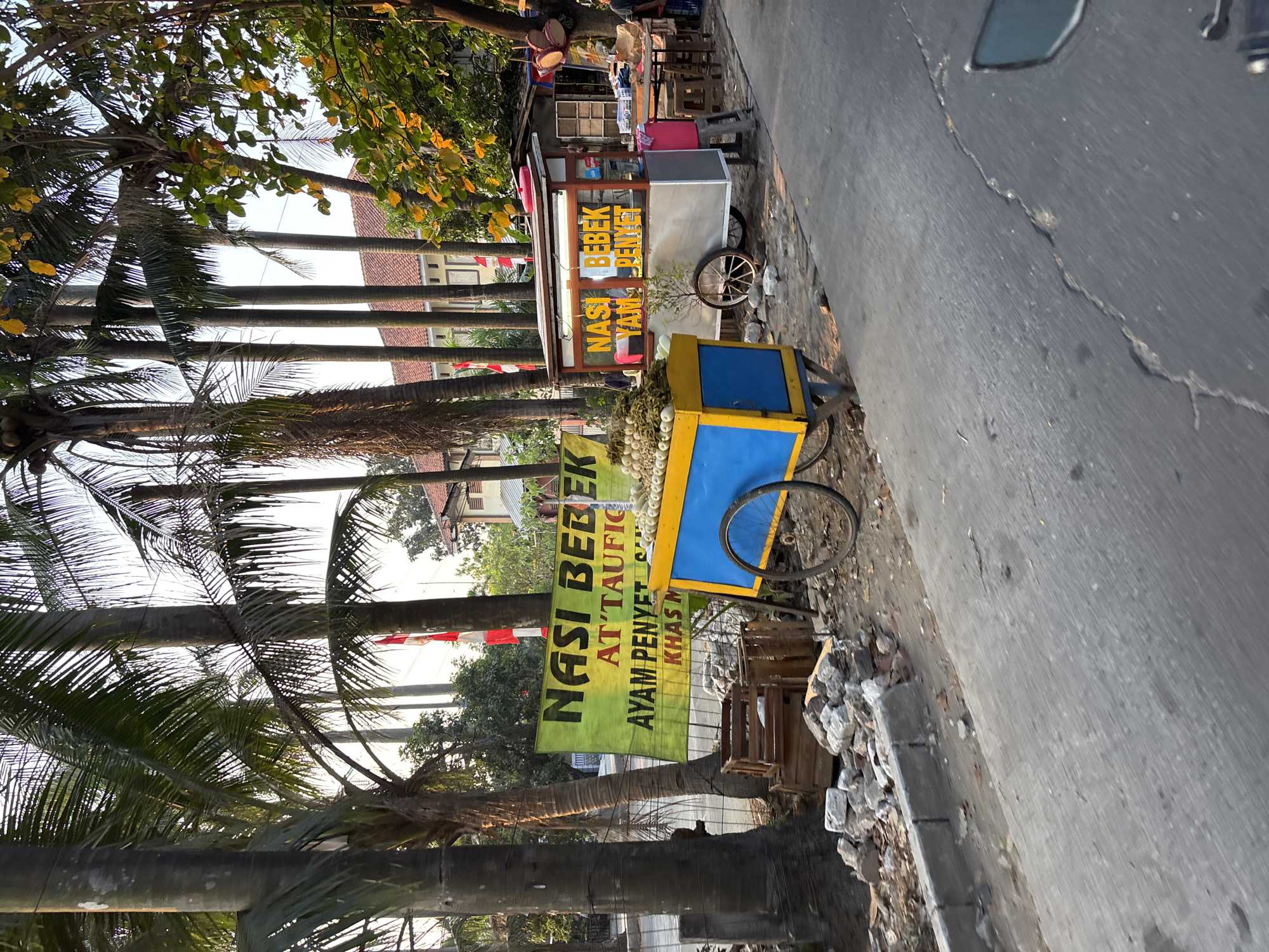 A street-level, medium shot captures a roadside food stall in Bekasi, Indonesia. The scene is dominated by tall, slender palm trees lining a moderately busy road. In the foreground, a blue and yellow food cart, piled high with what appears to be fried food items, sits on the side of the road. Behind it, a larger, silver food stall with a red roof displays the words "NASI BEBEK" (duck rice) and "AYAM PENYET" (smashed chicken) on its signage. A red and white Indonesian flag is visible on a pole behind the stalls. The background reveals a building with a tiled roof, suggesting a residential or commercial area. Debris, including broken bricks and wooden crates, litters the ground near the food carts, indicating ongoing construction or general disarray. The lighting suggests it is either early morning or late afternoon, with a warm, natural light illuminating the scene. No people are clearly visible in the frame, though the presence of the food stalls implies commercial activity.