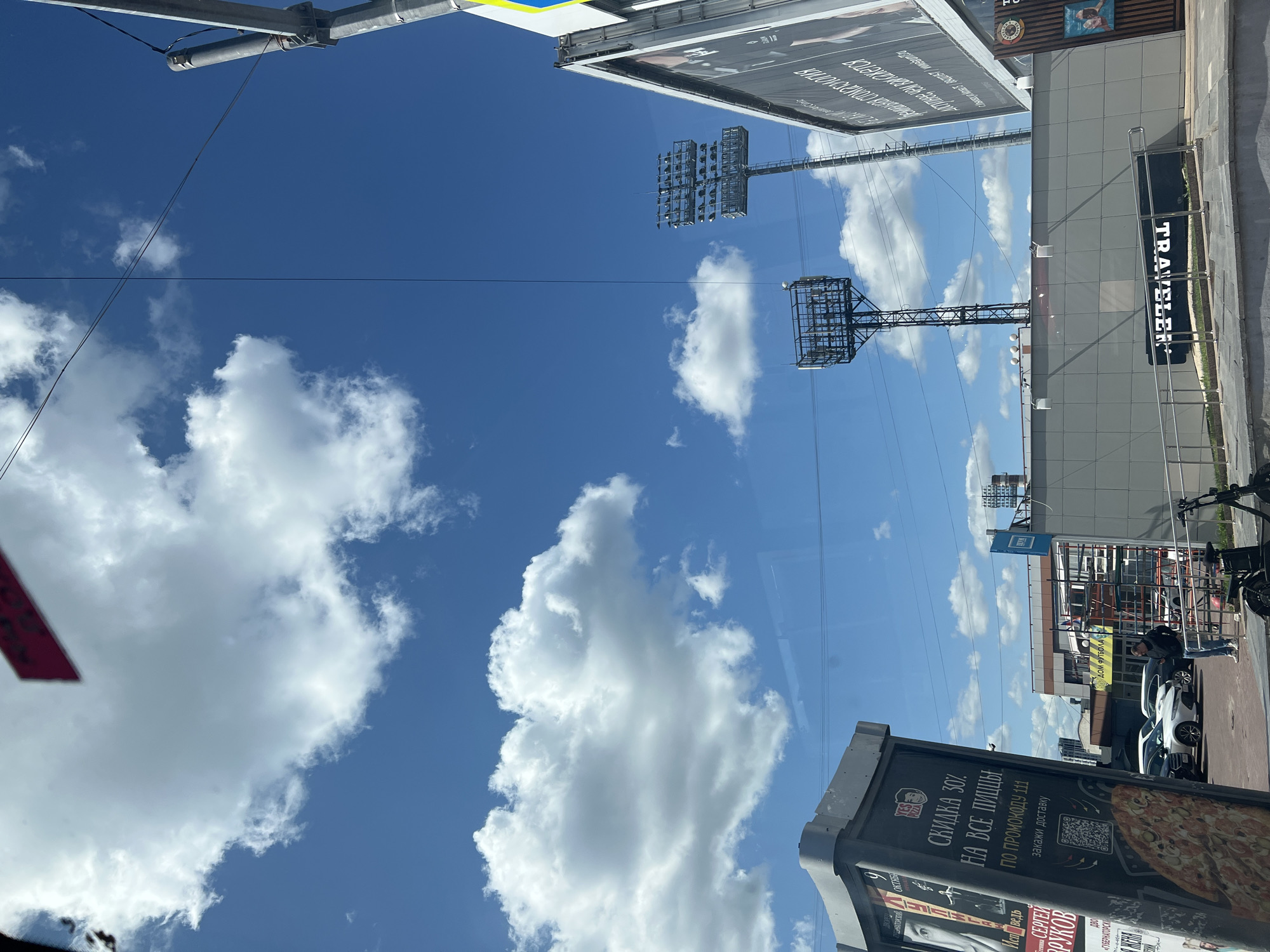The image shows an outdoor scene with a clear blue sky dotted with white cumulus clouds. In the foreground, on the left, is a tall, angled advertising kiosk with posters displaying images and text. Text visible on the kiosk includes "СКИДКА 30% НА ВСЕ ПИЦЦЫ" (30% discount on all pizzas) and a QR code. To the right of the kiosk, several cars are parked in a lot. In the mid-ground, there are two tall stadium lights with multiple lamps, one on the left and a larger one on the right, against the sky. Behind the stadium lights, a long, modern-looking building with a grey, tiled facade is visible. A sign with the word "TRAVELER" in large white letters is mounted on the facade. Further in the background, more buildings and structures are present. The overall scene suggests a public space, possibly near a sports venue or an urban area. Based on the Cyrillic text on the advertising kiosk, the location is likely in Russia. A person in a dark jacket and light-colored pants is standing in the parking lot. Another person is seen from the chest up, also in the parking lot. The image appears to be taken from inside a vehicle, as indicated by reflections on the surface.
This image is located in Ulyanovsk, Russia, specifically near the "Zheltoksiners" soccer stadium. The advertising kiosk is located at the entrance to the stadium, and the stadium lights are part of the sports facility.