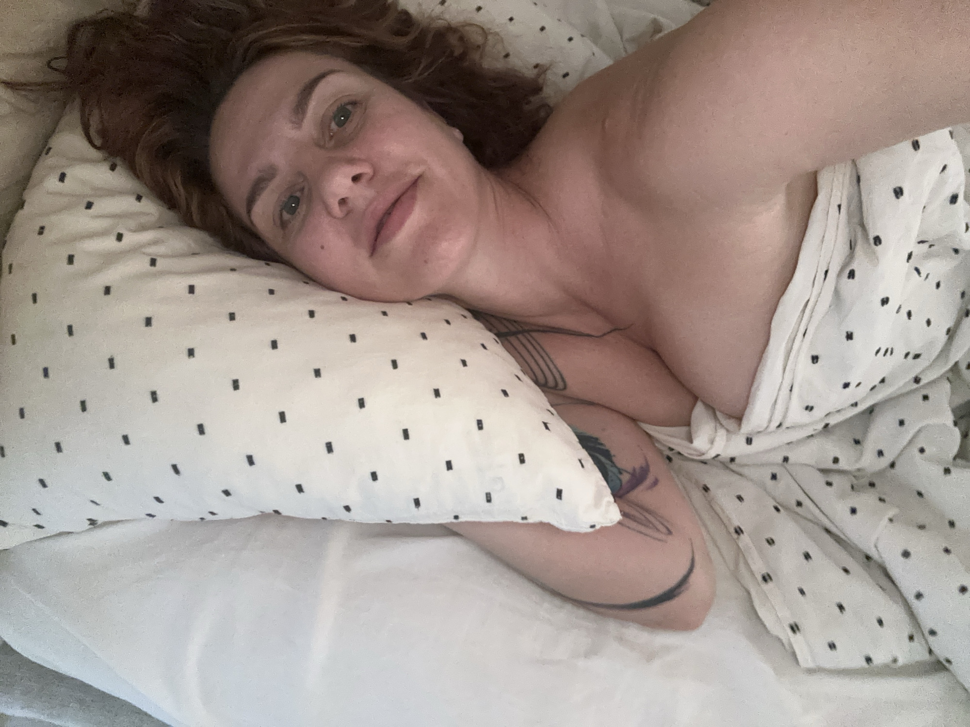 An adult female is captured in a selfie, lying in bed with a relaxed expression. She has reddish-brown, wavy hair spread around her head and clear blue eyes looking directly at the camera. Her face shows natural skin with no visible makeup. She is positioned on her right side, with her head resting on a white pillow adorned with small black square patterns. Her bare shoulders and upper chest are visible, partially covered by a white sheet or blanket with the same black square pattern. Several tattoos are visible on her left arm and shoulder area, including a colorful, possibly floral or abstract design on her upper arm and dark lines on her forearm. The setting appears to be a bedroom, with white bedding and natural light suggesting it is daytime, possibly morning. The overall atmosphere is intimate and serene. No text is visible within the image. The provided context indicates the location is Sochi, Russia.