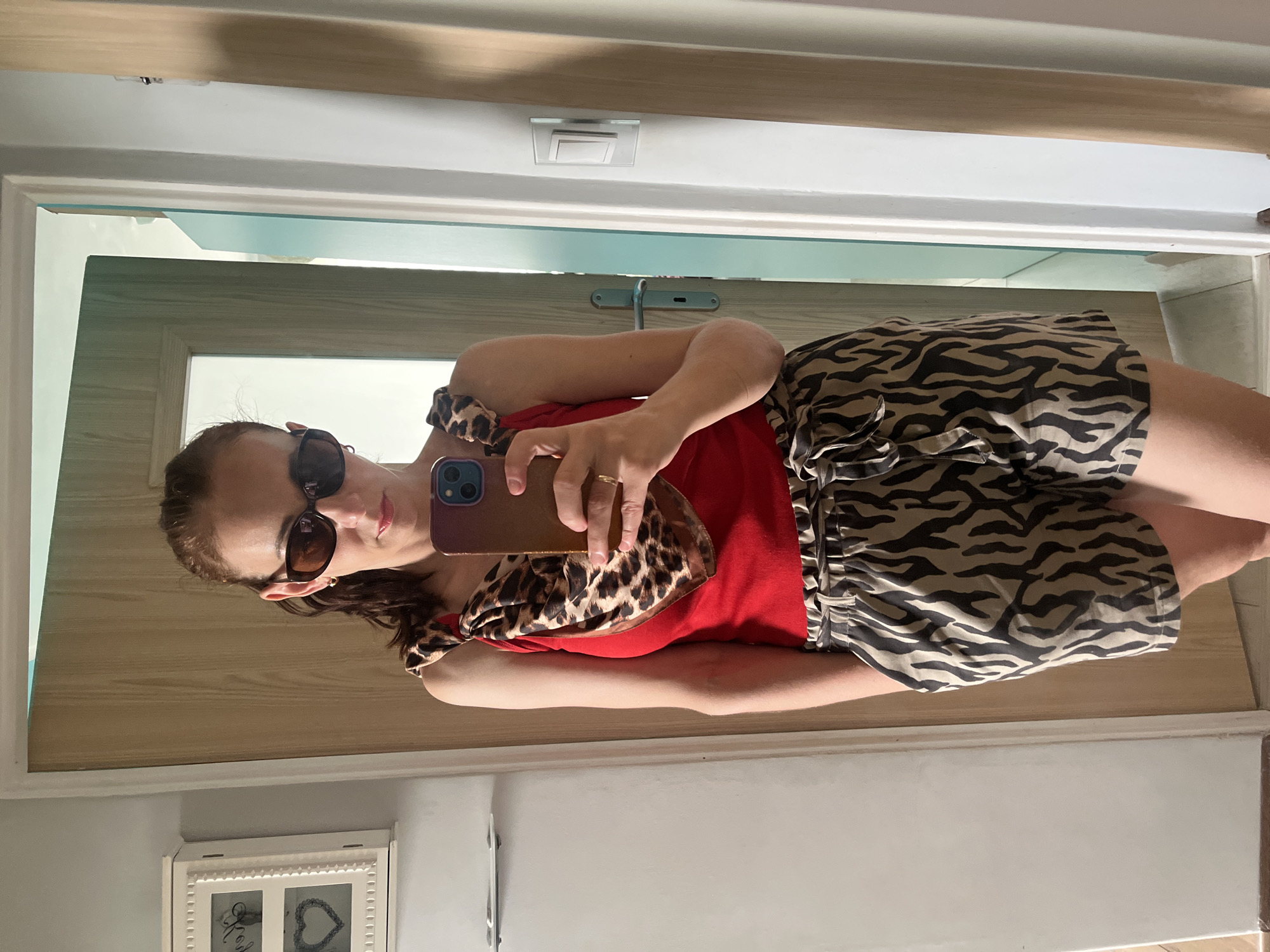 A mirror selfie captures a woman from the chest up, positioned horizontally in the image due to rotation. She holds a smartphone with a brown case in her left hand, extending it towards the mirror to take the photo. The woman is wearing a vibrant red top, zebra-print shorts, and a leopard-print scarf tied around her neck or shoulders. She sports dark sunglasses that obscure her eyes, and a gold-colored ring is visible on her left ring finger. Her lips show a neutral or slight smile. The background, reflected in the mirror, indicates an indoor setting featuring a light-colored wooden door frame, white wall trim, and a light blue wall or door. A white light switch fixture is visible on the ceiling above her reflection. In the lower left corner, a portion of a white framed picture rests on a gray wall, displaying two compartments, one with a cursive letter (possibly 'L' or 'E') and the other with a heart shape. The lighting appears bright, suggesting an indoor environment with natural light sources reflecting in the mirror. No explicit text is clearly legible other than the aforementioned cursive element and heart shape in the framed picture.