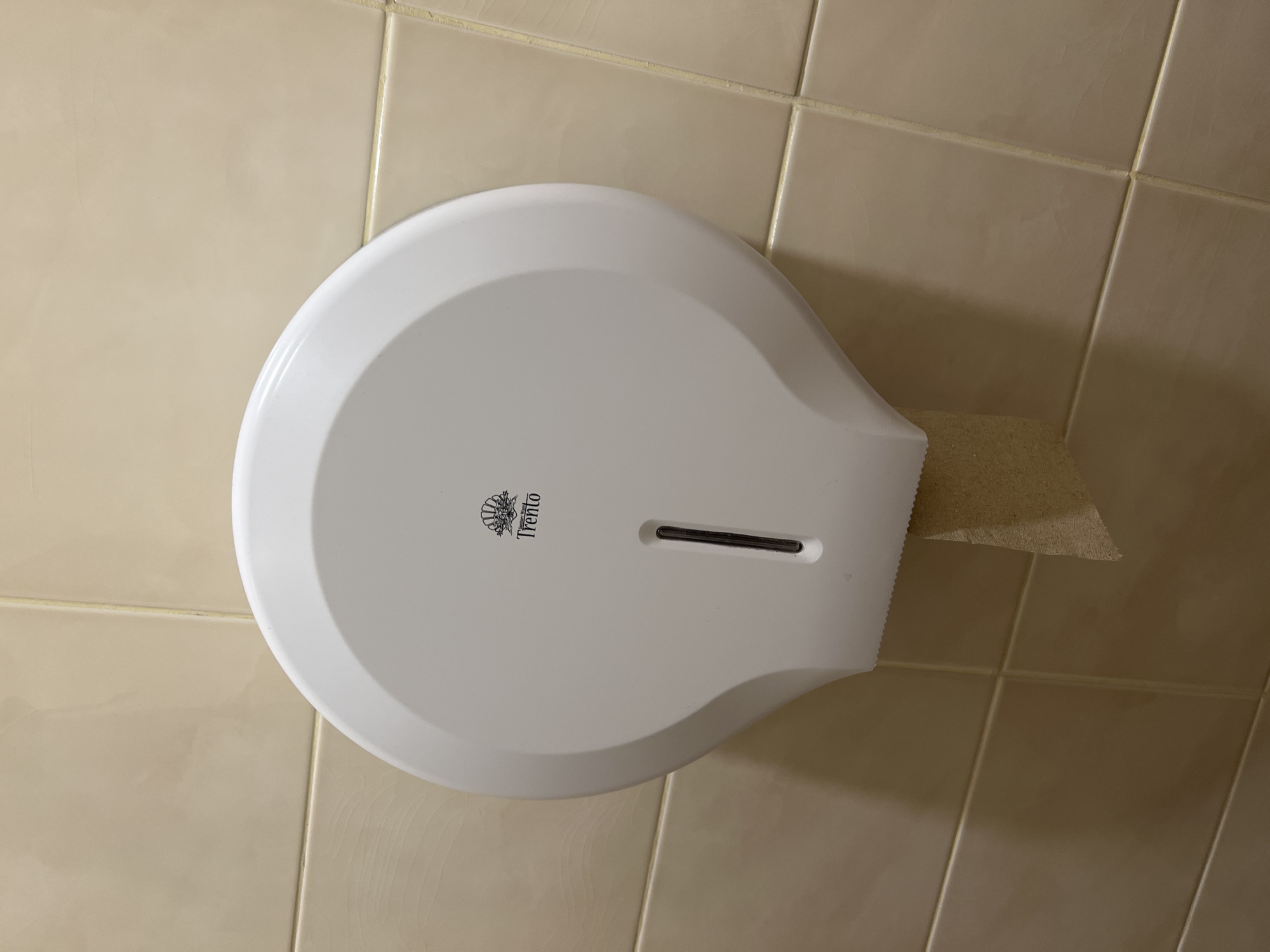 A white, wall-mounted paper towel dispenser, branded "Trento," is installed on a tiled wall. A paper towel protrudes from the dispenser. The tiles are light beige in color. The location and date are unknown.