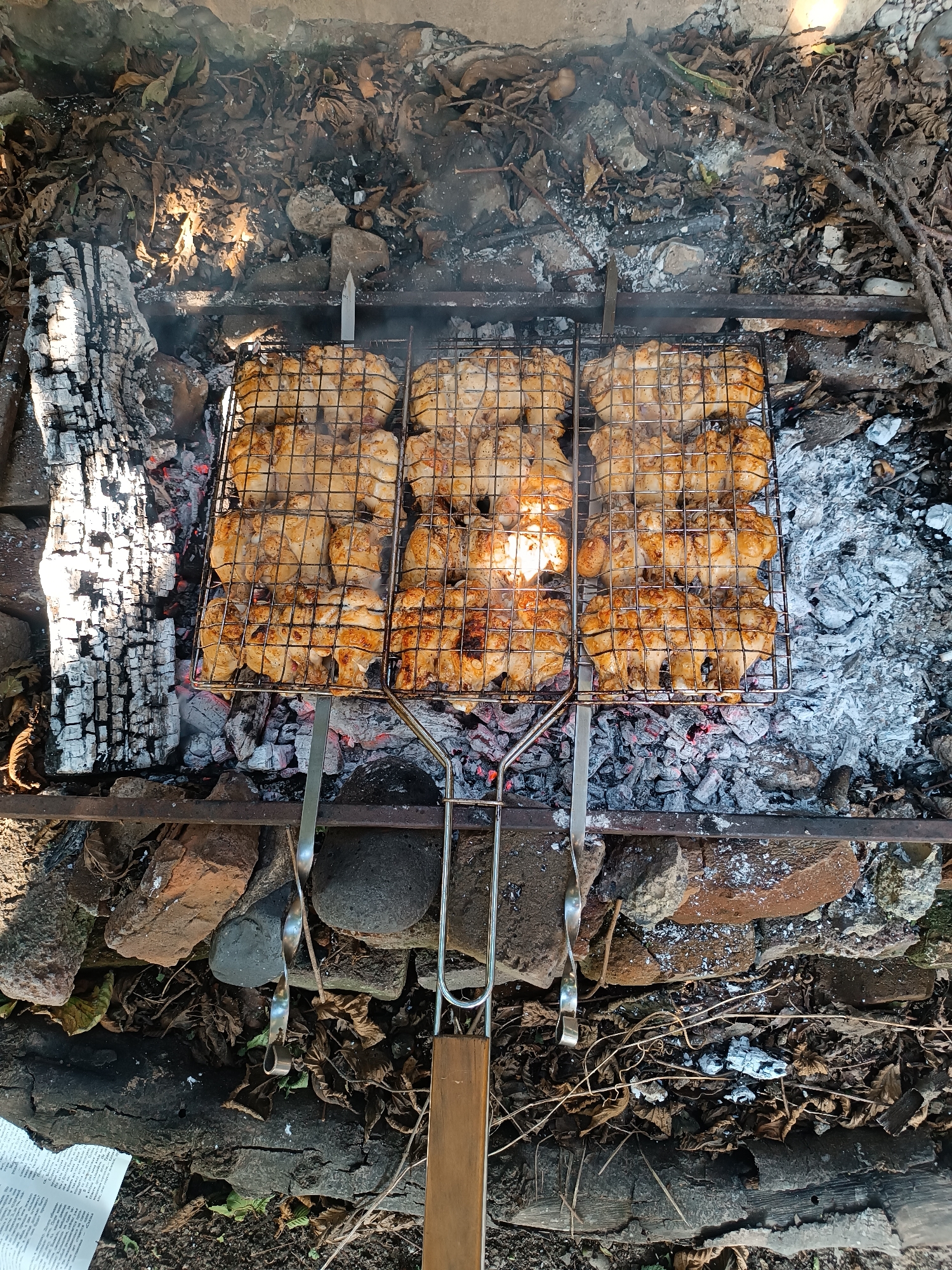 This is an overhead, eye-level shot of chicken pieces cooking on a grill over a fire. The chicken is golden brown and appears to be on a metal grate over hot coals and ash. The grill is placed outdoors, with burnt logs and dry leaves surrounding it, suggesting a natural or rustic setting. Some stones and brick fragments are also visible around the fire pit. The lighting suggests it is daytime, with sunlight casting shadows. The scene evokes a feeling of outdoor cooking and possibly a picnic or gathering. The location, Khashuri, Georgia, suggests a Georgian culinary context for the grilled chicken.